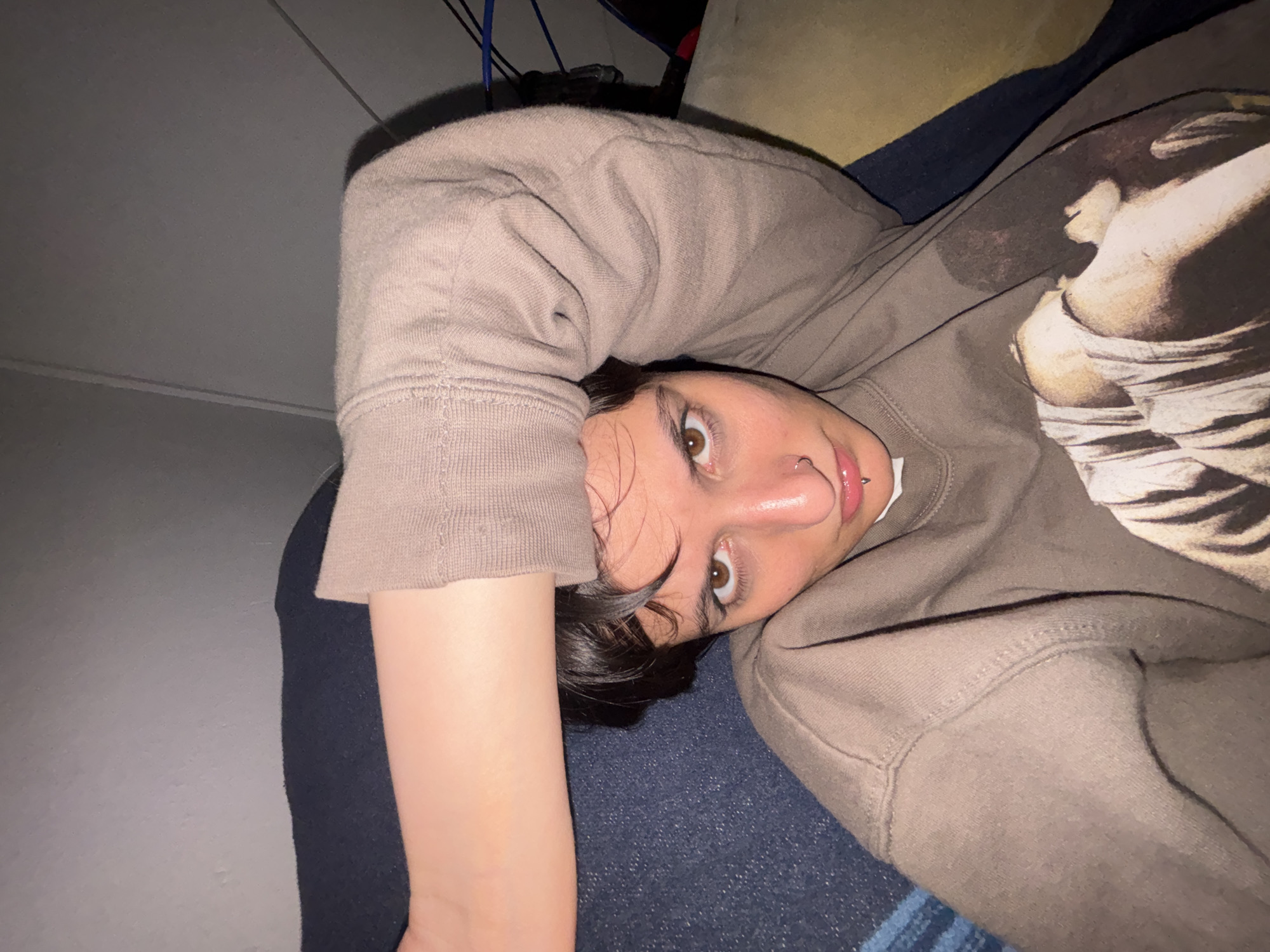 A close-up, angled shot shows a person lying down, possibly on a bed or a couch. The person has brown eyes, dark hair, and a piercing on their lip and nose. They are wearing a brown sweatshirt with what appears to be a faded design on it. The person's arm is extended towards the camera, partially obscuring the view. The background includes a wall and what appears to be bedding or cushions. The exact location in Sofia, Bulgaria, is not discernible from the image.