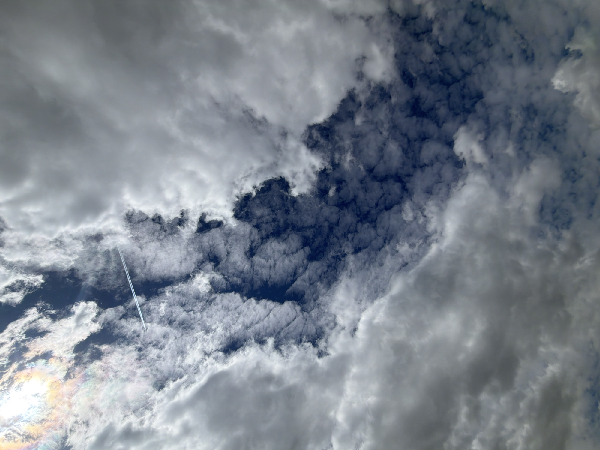The image shows a cloudy sky with patches of blue visible through the breaks in the clouds. A contrail from an airplane is visible in one of the blue patches. There is also an iridescent area possibly near the sun. The location is unknown and there is no indication of Tokkari-Lend, Russia. There are no people, subjects, objects, actions or interactions visible.