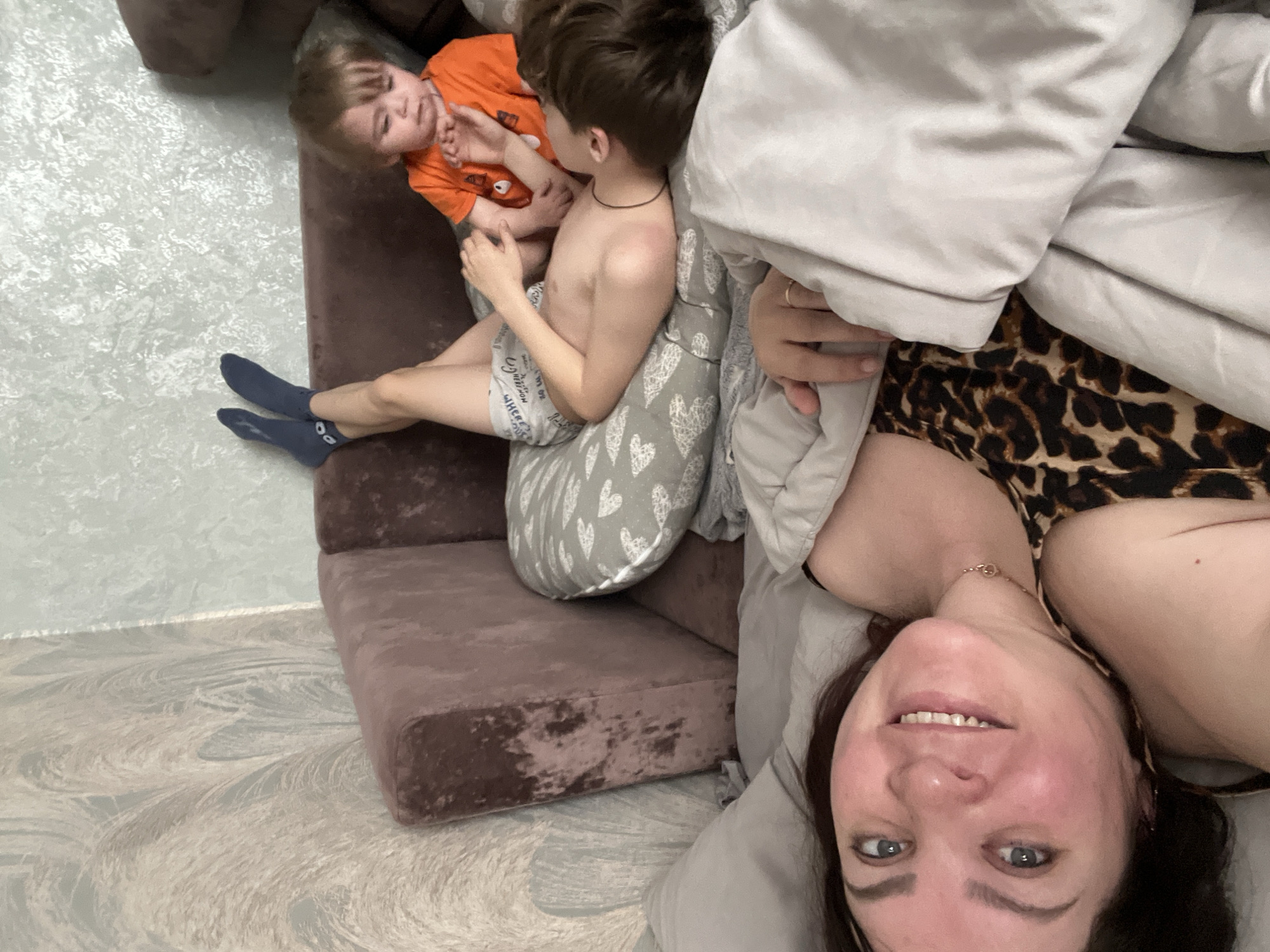 The image shows a woman lying on a bed or sofa with a leopard print top, covered partially by a light gray blanket or duvet. Her face is visible, positioned upside down relative to the scene, showing a smile.
Two young children are in the upper part of the image. One is wearing an orange shirt, and the other is shirtless, seated on what appears to be a brown velvet sofa or sectional piece with a patterned gray pillow behind them.
The floor is visible in the lower left corner and has a textured surface. The location is specified as Noviy, Russia, though the scene is clearly indoors. The interaction between the woman and children isn't explicitly clear but suggests a relaxed, domestic setting.