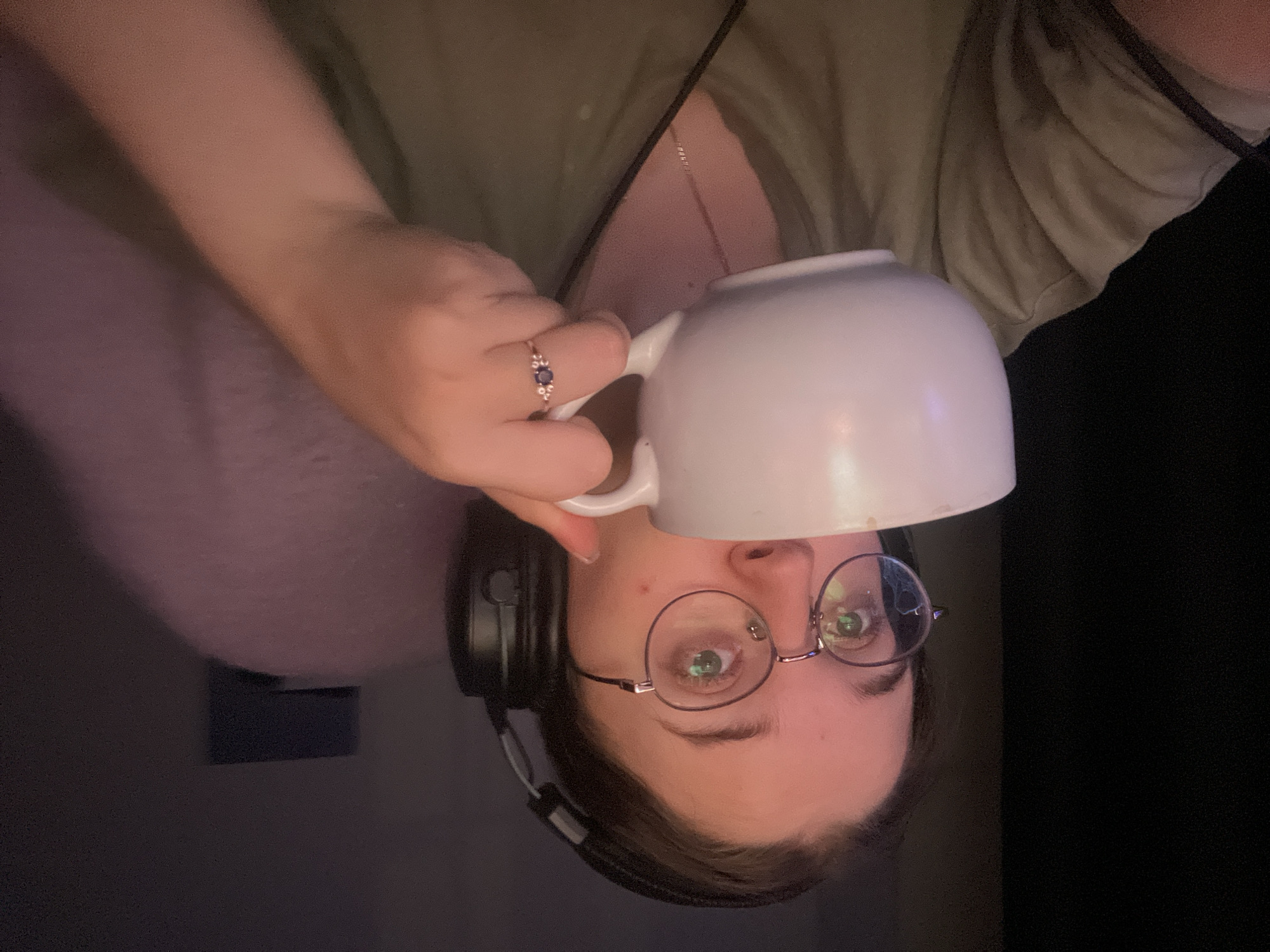 A person with glasses and headphones holds a white mug. The person wears a green shirt and a ring with a dark stone. The background is dark. The city location is not determinable from the image.