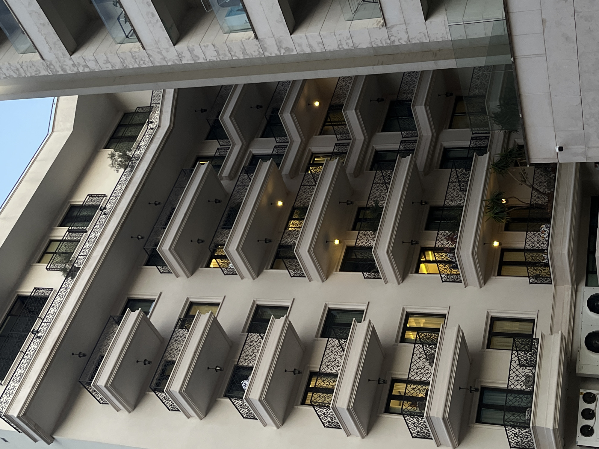 The image shows a close-up, eye-level, exterior shot of a modern, multi-story apartment building with multiple balconies. The building is primarily white with black wrought-iron railings on the balconies. Some balconies have plants and outdoor lighting visible. The building is located in Budva, Montenegro, as indicated by the presence of typical Mediterranean architectural elements, such as the style of the balconies and the building's facade. The neighboring building on the right appears to be constructed of lighter-colored stone with glass railings on its balconies. The sky visible at the top of the image is a pale blue, suggesting it is either early morning or late afternoon. There are no people or animals visible in the image. The focus is on the architectural details of the buildings.