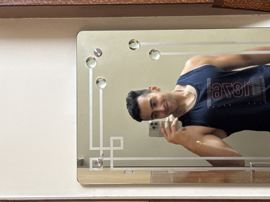 The image is a mirror selfie taken indoors by a man. The central focus is the man's reflection in a rectangular mirror, which is mounted on a plain, light-colored wall. A wooden frame is visible at the top and bottom edges of the overall image, bordering the wall space around the mirror.
The mirror itself has a distinctive design, featuring frosted or etched glass patterns. These include two vertical lines running down the left side, with circular, crystal-like embellishments or decorative screw covers placed at intervals along these lines and also across the top edge. There's also an intricate, angular geometric pattern in the lower-left corner of the mirrored surface.
The man reflected in the mirror has short, dark hair and is smiling faintly towards the camera of his smartphone, which he is holding up with his right hand to take the selfie. He is wearing a dark blue or black sleeveless tank top. On the front of his tank top, there is mirrored text that reads "ar8I" (which would be "18ra" or similar when un-mirrored) and some small dots below it. He also wears a silver chain around his neck and a thin gold bracelet on his right wrist. His pose suggests he is leaning slightly, and his upper body shows some muscle definition. The reflection appears clear and well-lit.