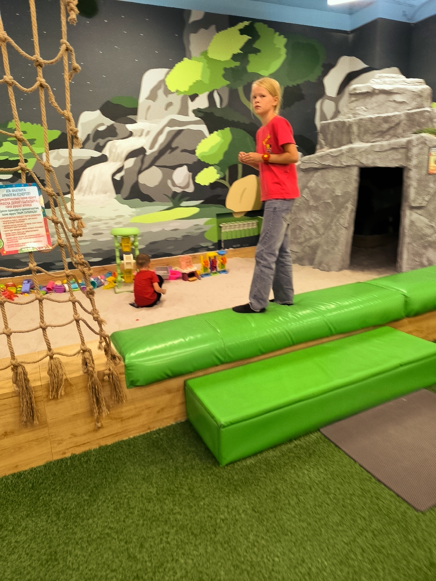 A young blonde girl in a red t-shirt and grey pants stands on a green padded beam in an indoor play area. To her left, a young boy in a red shirt is sitting on the sand. In the background, there is a mural of a waterfall and trees, and a rock-like structure. The floor is covered with artificial grass. A rope net hangs in the foreground. The location is identified as the city administration of Qarağandı, Kazakhstan.