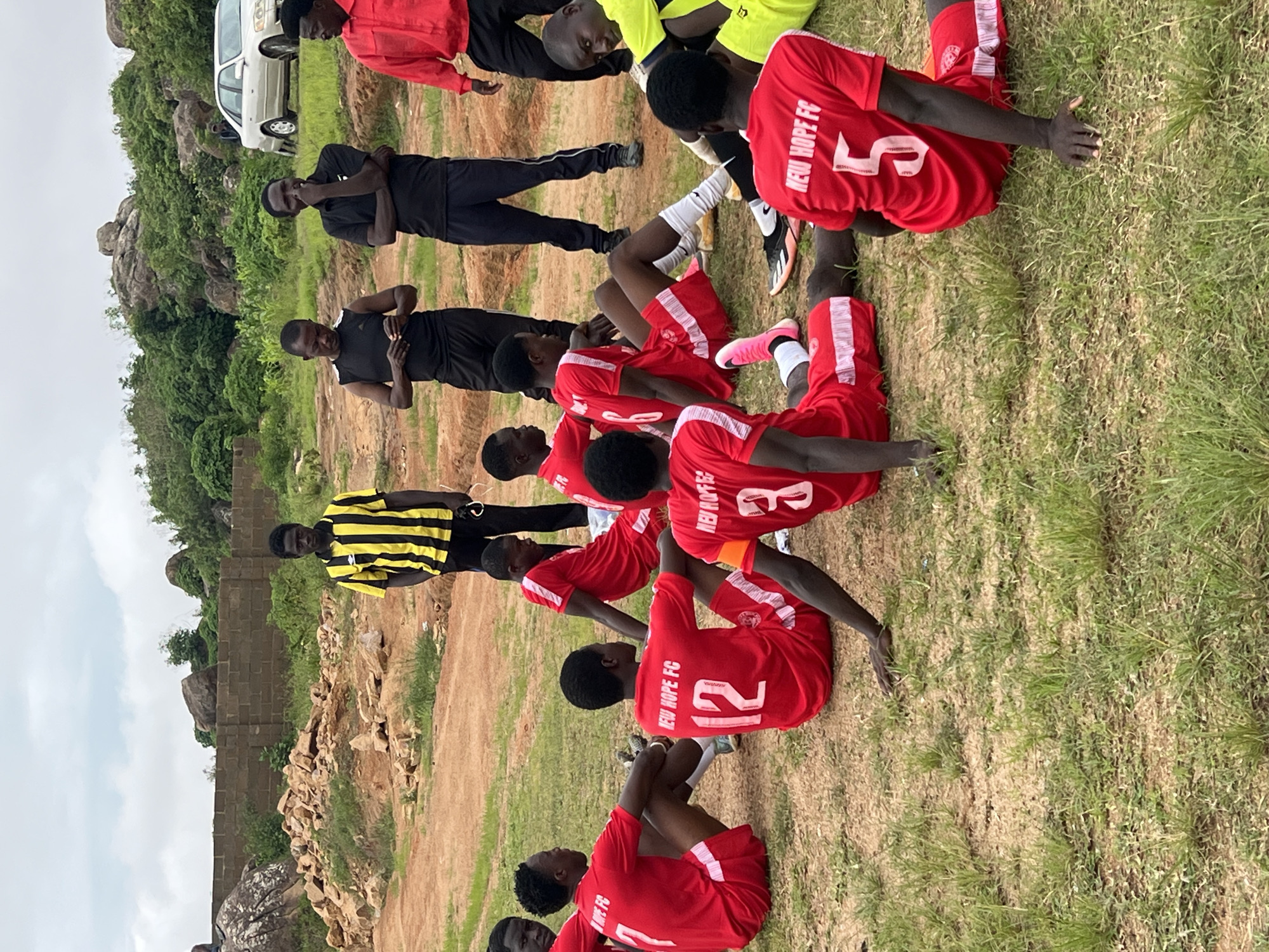 In the city of Jos, Nigeria, a group of young men, presumably a soccer team, are gathered on a grassy, uneven terrain. They are mostly wearing red soccer jerseys with white lettering and trim, some with numbers visible. Some are sitting on the ground, while others are standing. In the background, there is a wall made of brown bricks, a pile of rocks and dirt, lush green vegetation, and rocky hills under a cloudy sky. A silver car is parked in the distance. The overall scene suggests a training session or a break during a game in an outdoor, possibly rural, area on the outskirts of Jos.