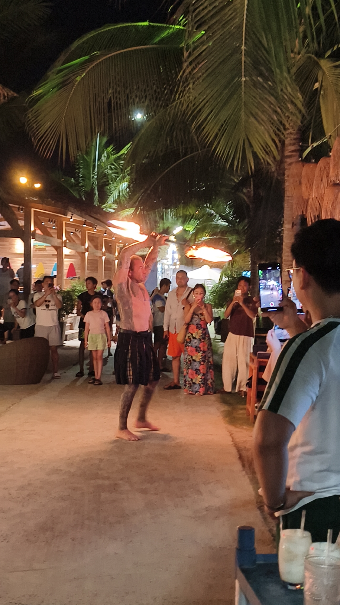 This image depicts a fire performance taking place at night, likely on a beach or outdoor venue in Nha Trang, Vietnam. The scene is illuminated by artificial lights and the flames from the performer's props, creating a vibrant and energetic atmosphere.
The central subject is a male fire performer, shirtless and displaying tattoos, wearing a kilt-like garment. He is actively engaged in spinning fire props, with flames extending from his hands. He appears to be the focal point of attention for the onlookers gathered around him.
A diverse group of people are observing the performance. Some are standing, watching the act, while others are capturing the moment with their mobile phones. There are adults and children present, suggesting a family-friendly event. The onlookers are dressed in casual, warm-weather clothing.
In the background, there are hints of a tropical setting with palm trees and what appears to be a beachfront establishment with wooden structures and thatched roofs. The lighting suggests it is evening or night. The ground is a sandy or paved surface.
Notable details include the bright flames, the tattoos on the performer, the various mobile phones being used to record the event, and the tropical foliage. The overall mood seems to be one of entertainment and enjoyment, with people taking pleasure in the spectacle. No visible text is discernible in the image.