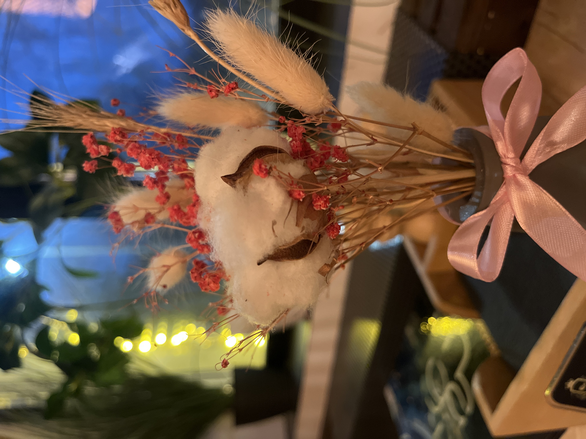 The image displays a close-up, vertically oriented shot of a decorative dried flower and cotton bouquet, set against a soft, out-of-focus background. The bouquet is the primary subject, held within a dark grey or black vase adorned with a delicate light pink satin ribbon tied in a bow.
The arrangement features two prominent fluffy white cotton bolls with visible brown husks. Interspersed among these are several creamy-white, elongated, fuzzy seed heads, likely bunny tail grass (Lagurus ovatus), alongside dried stalks of wheat or similar grasses. Small clusters of vibrant red, tiny dried flowers, possibly dried baby's breath, add a contrasting touch of color. The entire composition is built upon numerous thin, dry, light brown stems.
The background is heavily blurred, creating a bokeh effect. It includes sections of deep blue, possibly reflections or a blue surface, and several distinct circular yellow lights, suggesting artificial indoor lighting such as fairy lights or ambient reflections. Other blurred shapes in the background indicate an indoor setting, likely furniture or interior decor. No specific activity is occurring, as it is a static still life. The lighting suggests an evening or low-light time of day. No text is visible in the media file. The location context for this scene is Izhevsk, Russia.