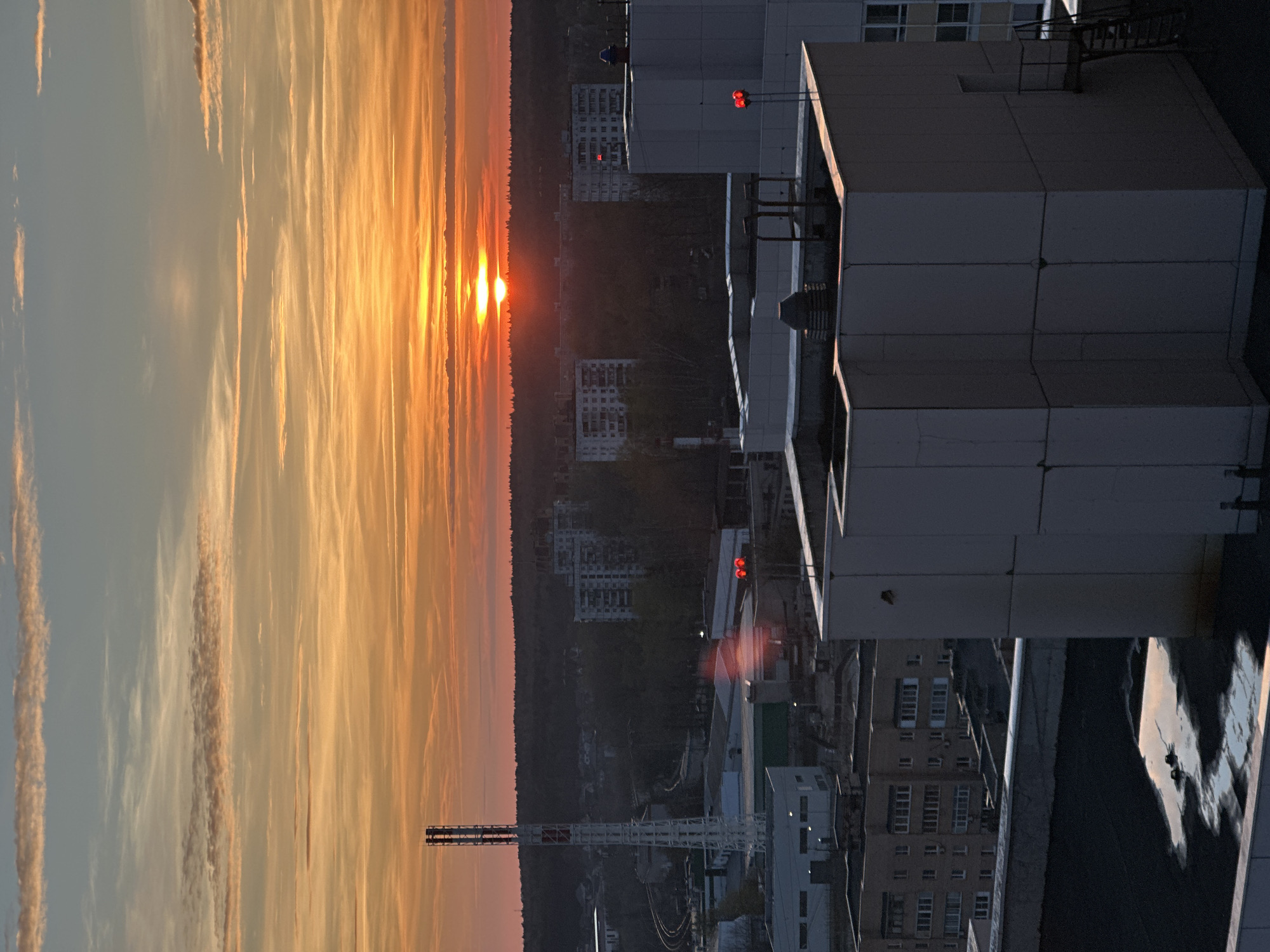 The image depicts an urban landscape at sunrise. The sun is positioned on the horizon, obscured by a treeline. The sky exhibits a gradient of colors, transitioning from orange near the horizon to a lighter hue higher up. In the foreground, a large building dominates the right side of the frame. Other buildings are visible further back, including one with red lights on top. A crane is present. The location is identified as the city of Krasnogorsk, Russia.