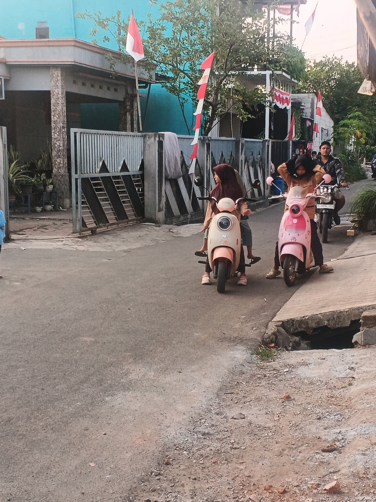 A group of young people are riding motorbikes down a paved street in what appears to be the city of Kadu, Indonesia. Two young women are riding in tandem on a beige scooter in the foreground, with the driver wearing a maroon hijab and the passenger a light brown headscarf. To their right, another young woman wearing a light brown hijab and matching outfit is riding a pink scooter, with the driver's hands positioned on the handlebars. Behind them, a young man in a camouflage shirt is riding a motorcycle. The street is lined with a metal gate and a house with a blue wall. Indonesian flags are visible, suggesting a patriotic holiday or celebration. The scene is set in a residential area with a mixture of houses and greenery.