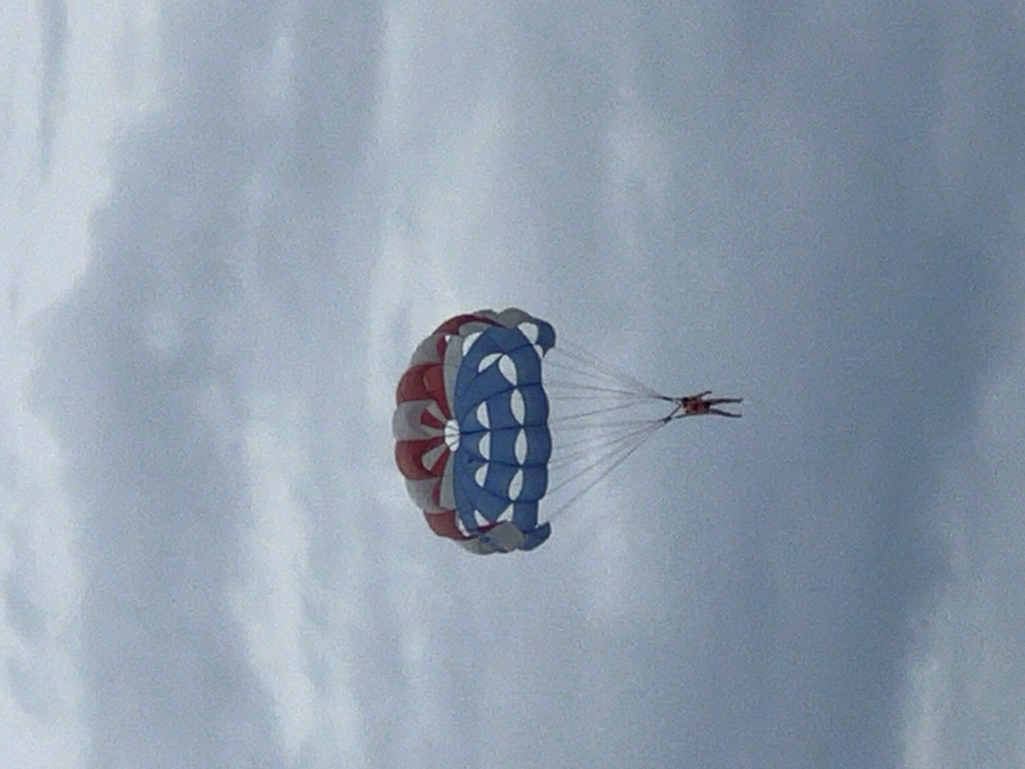 A paraglider with a red, white, and blue canopy is in mid-air against a cloudy sky, likely over the waters of Đà Nẵng, Vietnam. A person, wearing an orange vest and dark pants, is suspended beneath the canopy. The paraglider is positioned slightly to the left of the center of the frame. The surrounding environment is dominated by a vast expanse of gray, overcast clouds, suggesting a potentially overcast or misty day. The image captures a moment of recreational activity against a muted atmospheric backdrop.