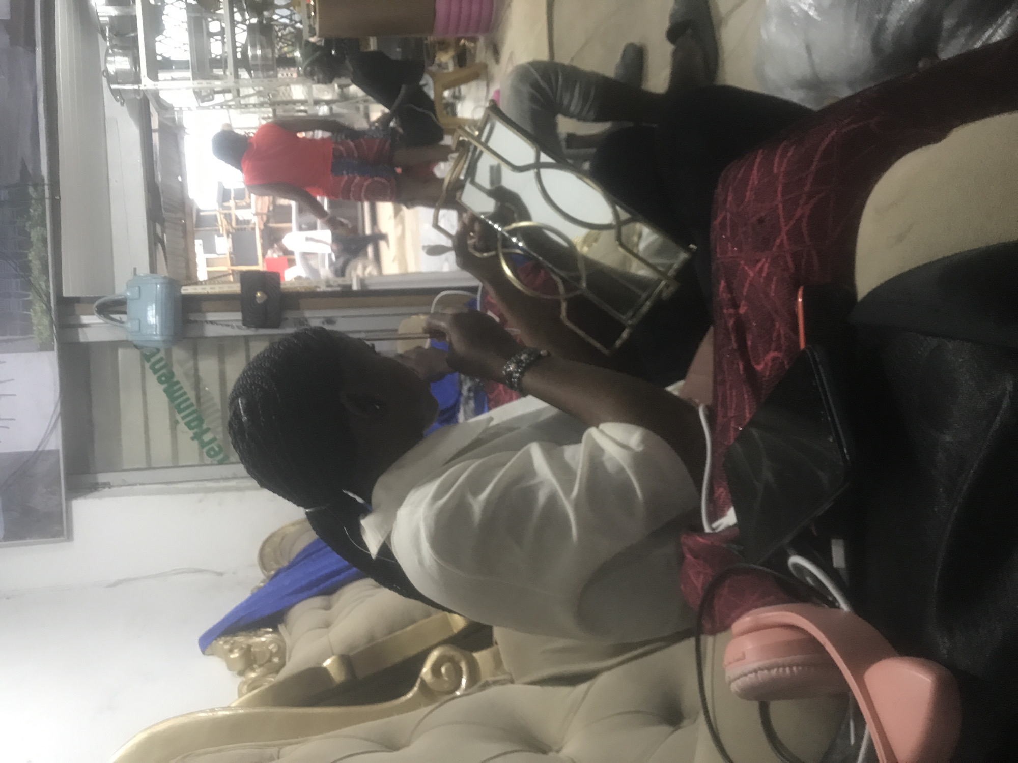 The image, oriented sideways, captures an indoor scene with several individuals and furnishings. In the immediate foreground, a person with braided black hair, wearing a white shirt, is seated on a large, ornate, light-colored tufted armchair or couch with golden decorative accents. This individual is holding a decorative mirror with an intricate gold frame and appears to be looking into it or manipulating something in front of it, with their hands raised. A bracelet is visible on their right wrist.
To the right of this person, on the armchair, are pink over-ear headphones, a black bag or jacket, and what appears to be a smartphone.
In the background, through what looks like a doorway or large window, another person wearing a red shirt and patterned shorts is visible, standing or walking. Farther back, another individual is partially visible. The background also reveals shelving or racks, suggesting a commercial or retail environment, possibly a salon, shop, or waiting area.
Other objects in the scene include a light blue handbag and a black handbag visible near the foreground. The lighting appears to be a mix of natural light from the background and indoor artificial lighting. Part of a word, "erainment" (likely from "entertainment"), is visible on a sign or glass panel in the background. The overall atmosphere seems casual.
