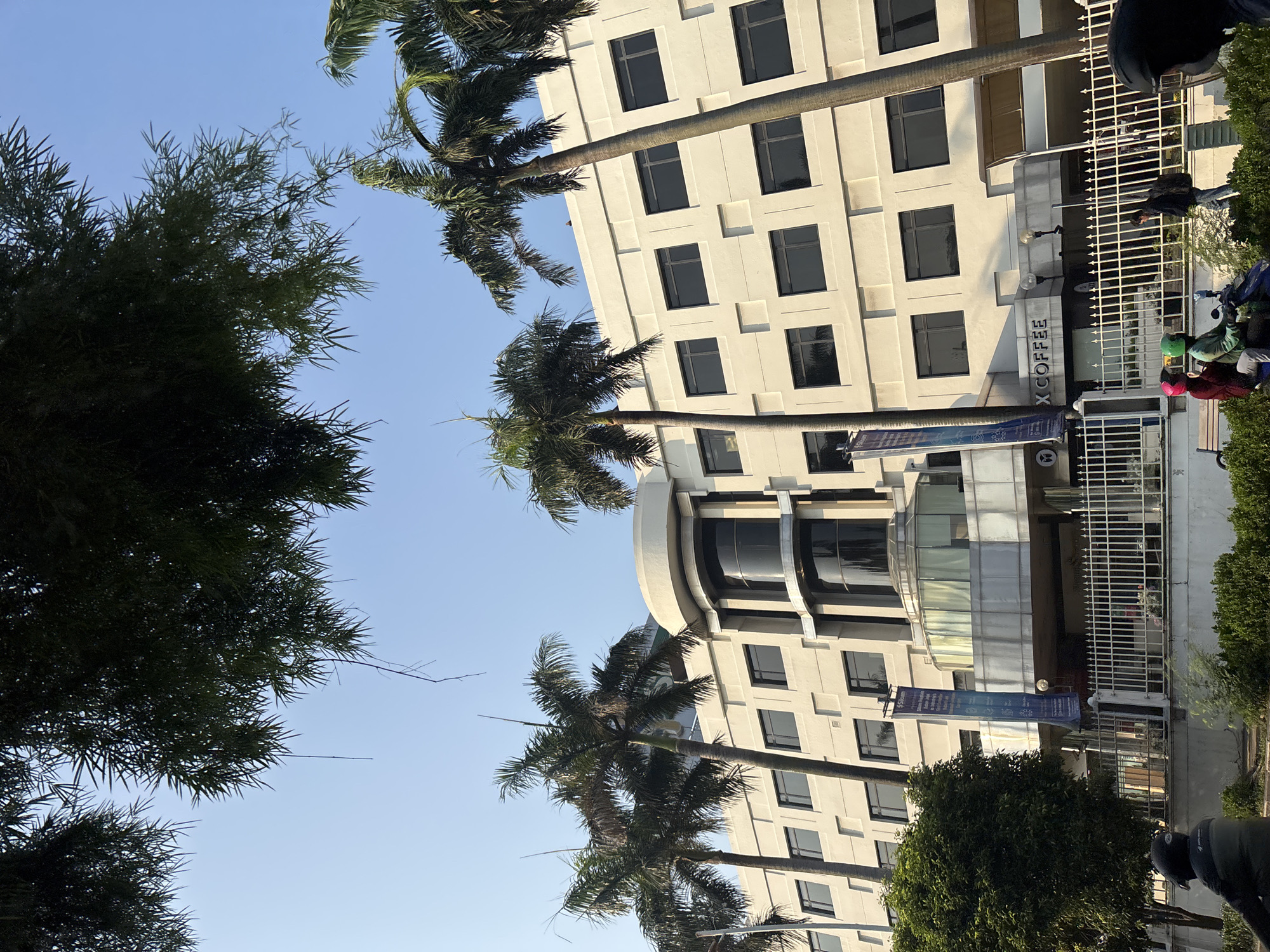 The image depicts the exterior of a hotel or office building, likely in West Jakarta, Indonesia, based on the architectural style and presence of palm trees. The building is light-colored with multiple floors, featuring many rectangular, dark-tinted windows. A prominent curved architectural element is visible on the upper floors. In front of the building, there is a white metal fence. Signs are attached to the building, including one vertical blue banner and another sign that reads "X COFFEE". Several palm trees are positioned in front of the building, with lush green foliage of what appears to be bamboo trees overhanging from the top of the frame. The sky is clear and blue, suggesting daytime. In the foreground, a person on a motorcycle is partially visible on the left, and two people on a motorcycle are visible on the right, with one wearing a red helmet and the other a green helmet. Another person is walking near the fence. The overall scene suggests a sunny day in an urban environment.