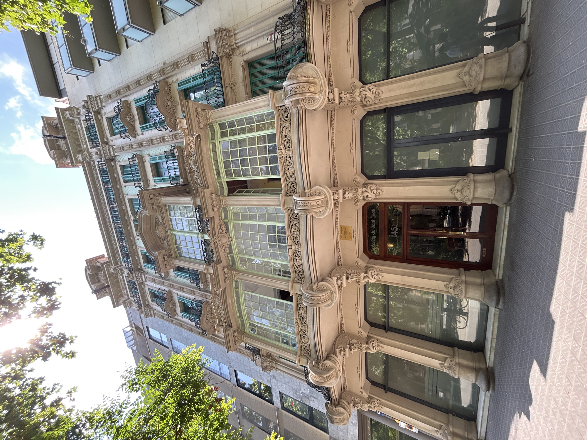 The image shows the facade of a building in Barcelona, Spain, with ornate architectural details in a Modernista style. The building is several stories tall, with multiple windows and balconies. The windows are divided into small panes and surrounded by decorative moldings. The balconies have wrought-iron railings. The building's color scheme consists of light browns, creams, and touches of green. A sign next to the building's entrance reads "Passatge de Sant Joan 145." The image is taken from a low angle, looking up at the building, with trees in the foreground partially obscuring the view. The pavement is visible in the foreground, with its patterned tiles. The building's architecture is the main focus.