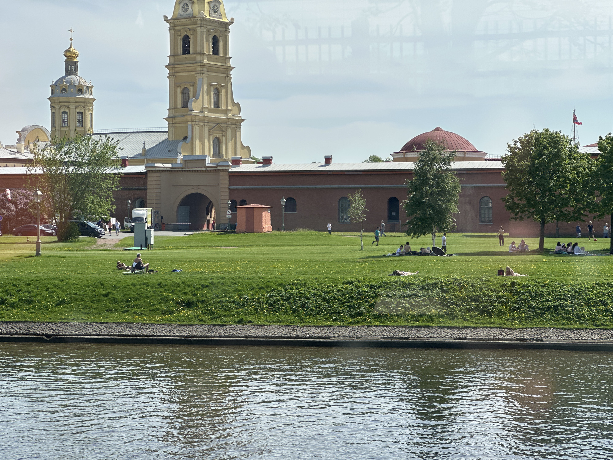 The image shows a view across a river towards a grassy area with several people relaxing on the lawn in Saint Petersburg, Russia. In the background stands the Peter and Paul Fortress, distinguished by its tall, yellow bell tower and a dome-topped church structure to the left. A red-brick building stretches across the mid-ground behind the lawn. People are scattered across the grass, some lying down and others sitting or standing. The river dominates the foreground with rippled water. A stone embankment separates the water from the grass. Reflections of the sky and surrounding structures are visible on the river’s surface. The sky is overcast with some areas appearing lighter.