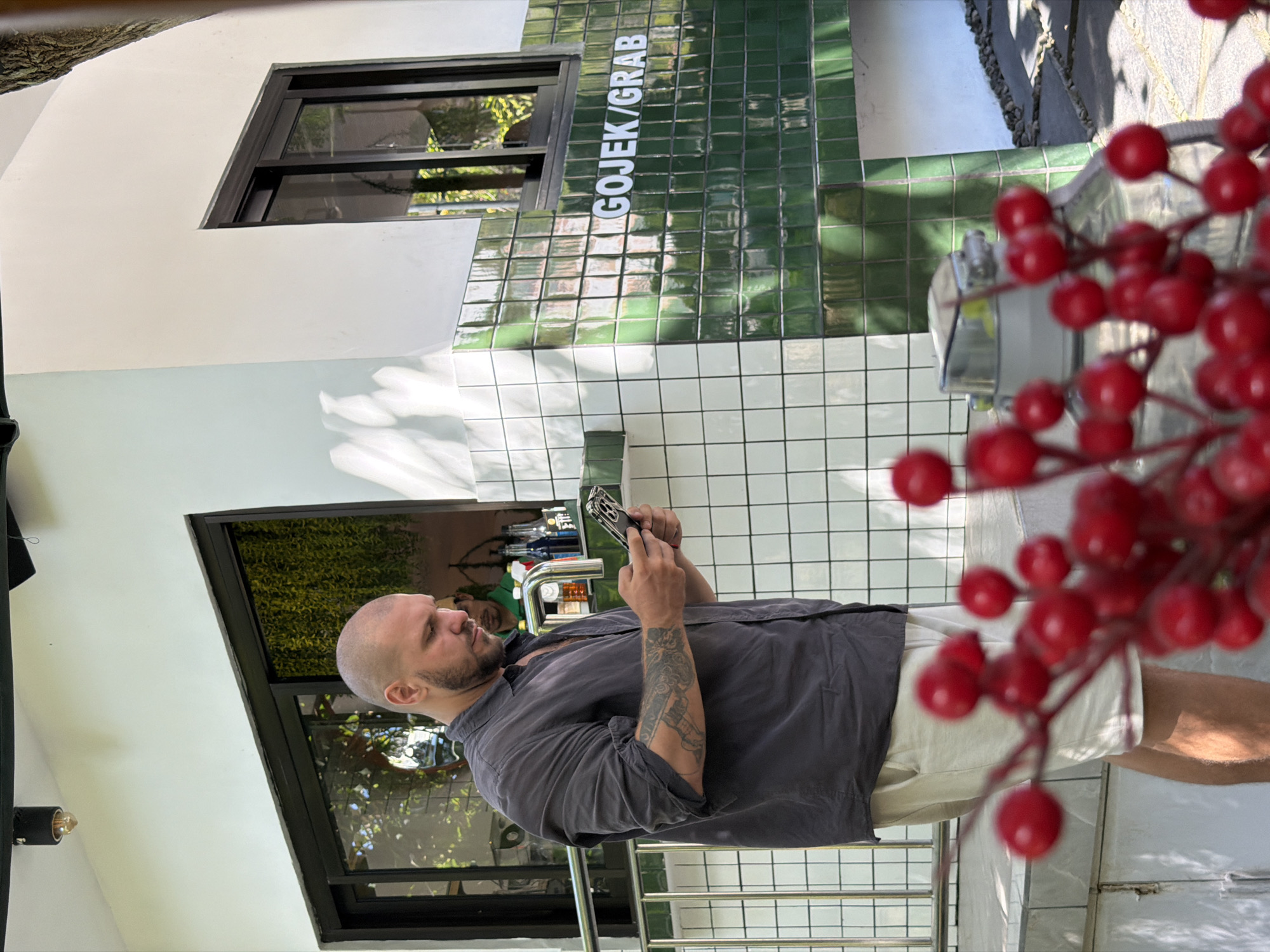 A bald, tattooed man in a dark grey, unbuttoned shirt and light beige shorts stands outside a building, looking at his phone. He is positioned in front of a white tiled wall with green tiled accents. To his right, the building features a prominent sign that reads "GOJEK/GRAB" in white lettering against the green tiles. Through a window behind him, another person in a green shirt can be seen. In the foreground, out of focus, are bright red berries on a stem. The building appears to be a commercial establishment, possibly a shop or service point, located in Kuta Selatan, Indonesia. The lighting suggests it is daytime.