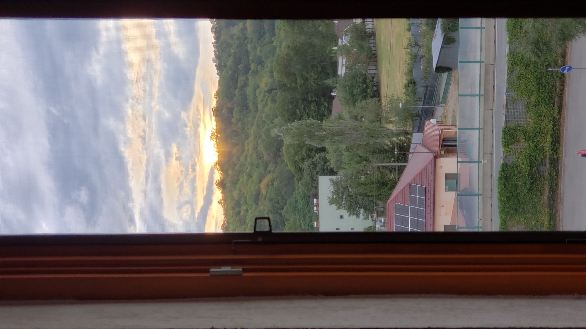 The image shows a view through an open wooden window frame, looking out onto a landscape. In the foreground, to the left, is the edge of a white textured wall and the wooden frame of the window. The view beyond the window reveals a suburban or semi-rural scene with buildings, trees, and a hill in the background.
In the middle ground, there is a building with a red roof covered in solar panels. Adjacent to it is a smaller building with a similar red roof. Further back, a taller, lighter-colored building is visible, surrounded by trees. A tall, slender tree stands prominently in front of this building. There is a road or path in front of the buildings, bordered by a green fence. Beyond this, there is a large expanse of green grass, possibly a field or park, with more trees and fences.
The background is dominated by a densely forested hill, with the setting sun peeking through the trees near the top, casting a warm glow and rays of light. The sky above is partly cloudy, with a mix of light and dark grey clouds, some of which are illuminated by the setting sun. The overall impression is a peaceful evening scene.
Based on the visual cues and a general knowledge of the landscape, this scene is likely located in Prešov, Slovakia. The combination of residential buildings, a nearby hill covered in trees, and the characteristic European style of architecture visible in the buildings suggests this location. The presence of solar panels on the roof also points to a modern development within a city.