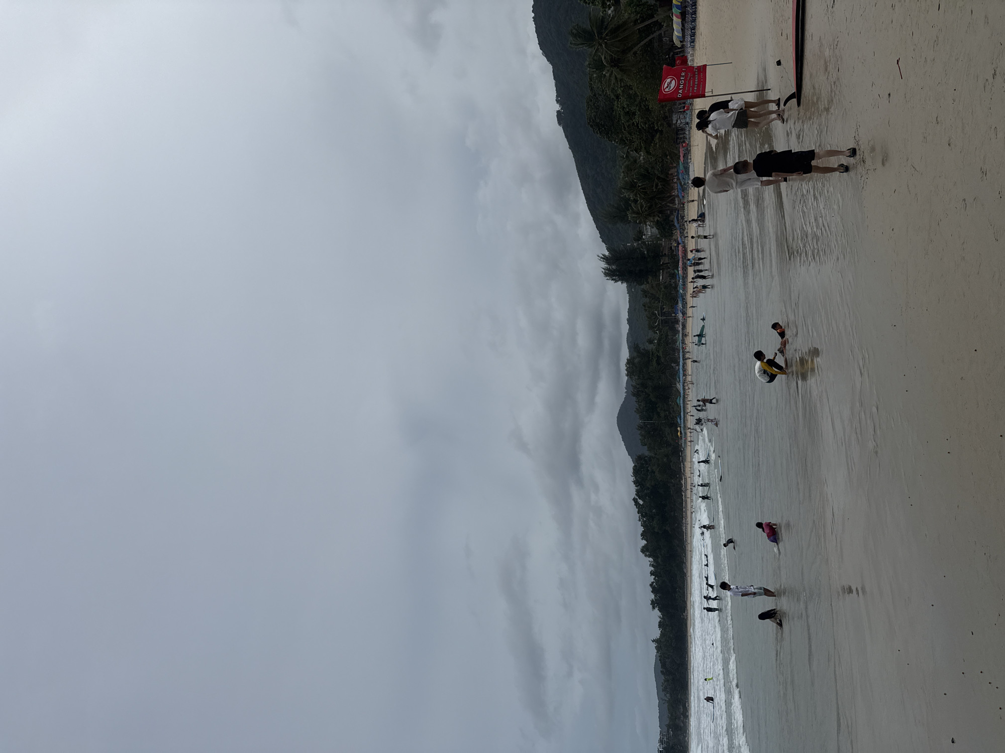 An overcast day at Karon Beach in Karon, Thailand, features a wide expanse of sandy beach with the ocean in the foreground and a tree-covered hillside in the background. The sea is calm with gentle waves. Several people are visible on the beach and in the shallow water. Some are wading or playing in the water, while others are walking along the shore or standing. A few individuals are in the ocean, including some on surfboards. A red flag with a white symbol is planted in the sand near the water's edge, accompanied by text in an unfamiliar language, likely indicating a warning or safety information. Beach umbrellas and lounge chairs are visible in the distance along the treeline. The overall atmosphere is one of a moderately populated beach on a cloudy day.