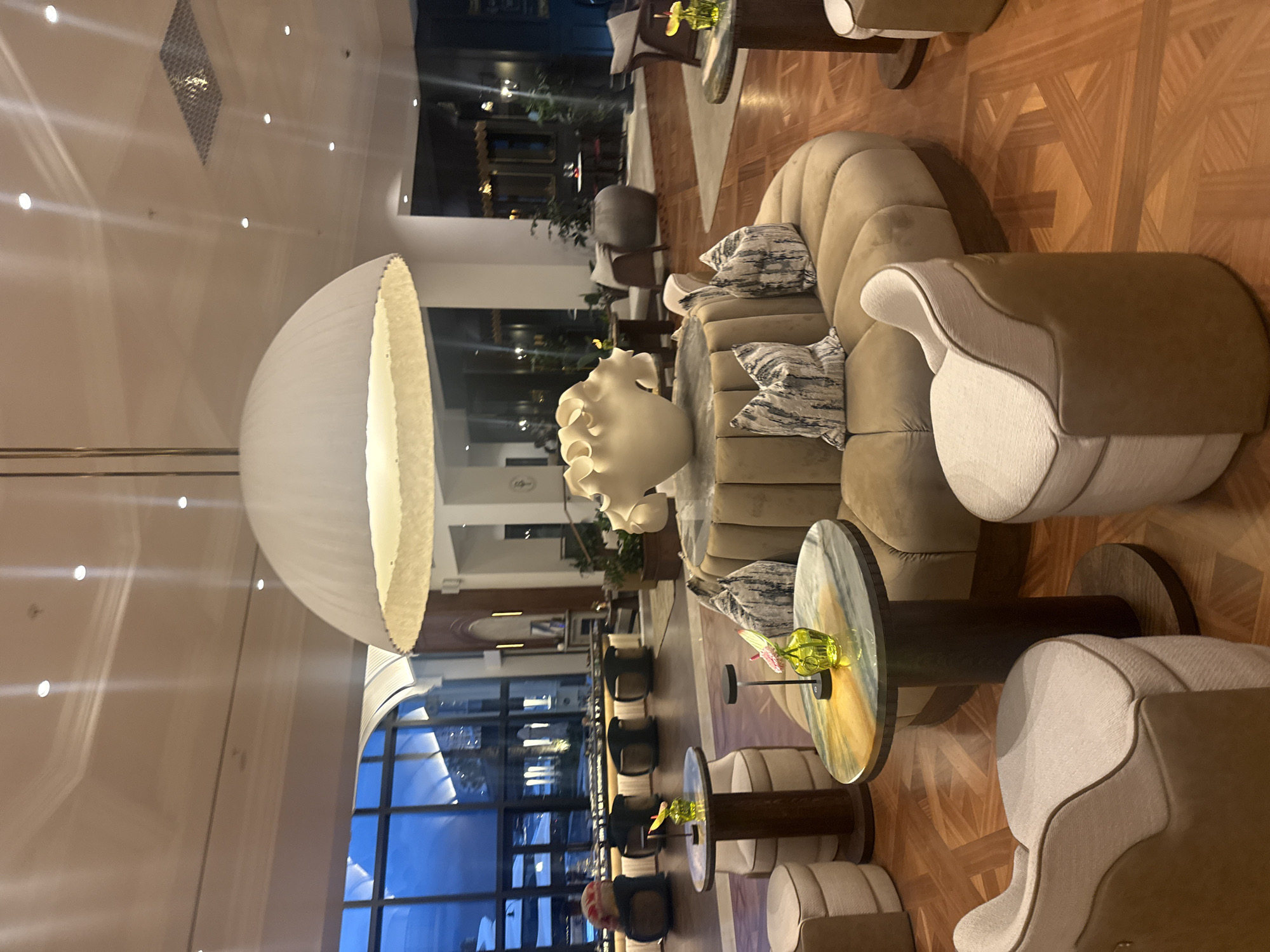 The image depicts the interior of a luxurious lobby or lounge area, likely in a hotel. The location is potentially within Cape Town, South Africa. The scene includes plush seating arrangements with a large, abstract, white sculpture serving as a centerpiece near several curved sofas with decorative pillows. There are small, round tables with glass tops and decorative arrangements, possibly flowers. The flooring is wooden with a parquet pattern. The space is well-lit, with a prominent overhead light fixture resembling an inverted dome. Tall windows offer an outside view, suggesting a location with visibility to the exterior. No people are clearly visible in the foreground, though there is a hint of figures and furniture in the background. The dominant colors are beige, white, and shades of brown, creating a warm and inviting atmosphere.