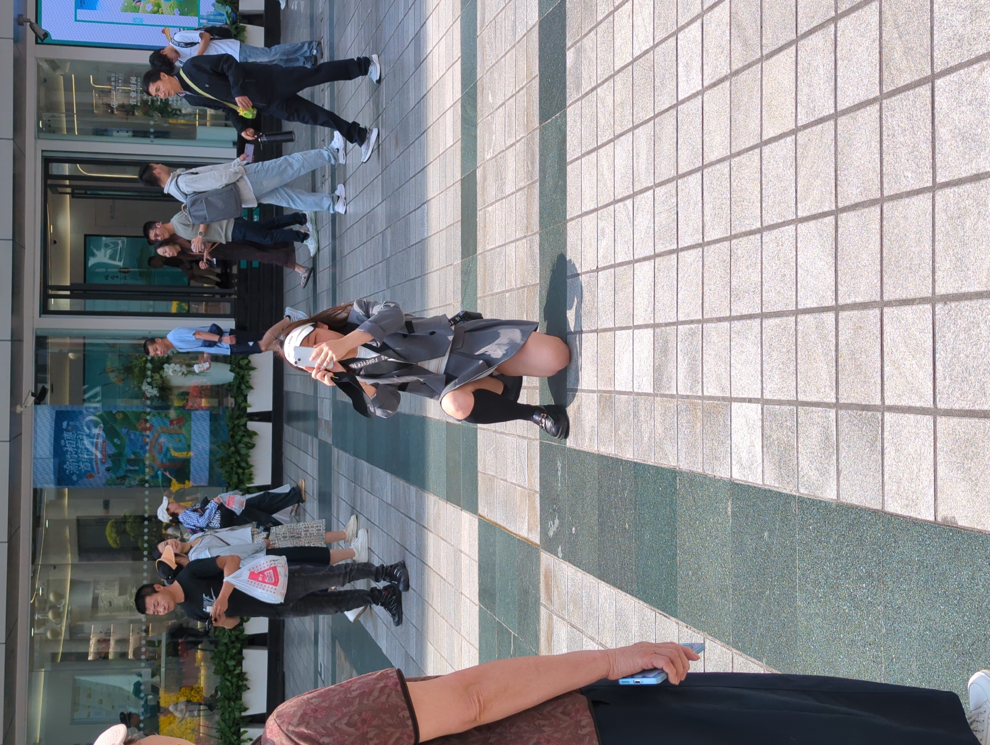 The photo captures a street scene in the Yuzhong District of China, revealing a series of individuals amidst a commercial or public space. In the foreground, an individual's arm and hand are holding a blue smartphone. A woman wearing a grey blazer, skirt and knee high socks, is positioned kneeling, using her phone to take photos or videos. The scene is composed of tiled flooring of grey and green with shadows that give it a sense of depth. Further away, a mix of pedestrians is observed. Some are walking, and appear to be looking into store windows. The backdrop includes storefronts with signage, posters, and greenery, creating a blend of commercial and natural elements.