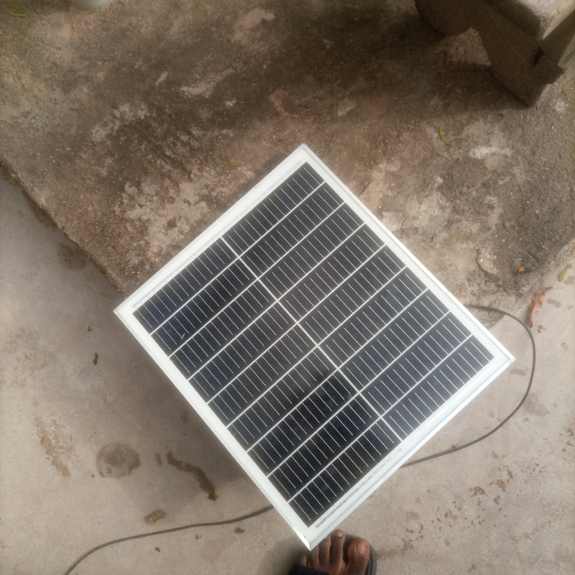 A solar panel with a white frame sits on a mottled gray concrete surface. The panel is oriented diagonally, with its top-left corner pointing towards the upper left of the frame. The surface of the solar panel is composed of numerous dark rectangular cells, divided by thin white lines. A thin black cable extends from the lower right corner of the solar panel and curves across the concrete. A person's foot, wearing dark footwear, is visible in the lower right portion of the image. The concrete surface appears worn and has irregular lighter and darker patches, suggesting it is outdoors and exposed to the elements. In the upper right of the image, a portion of a rough, textured object, possibly a block of stone or concrete, is visible. The background consists of the concrete ground, with some leaf litter scattered in places.