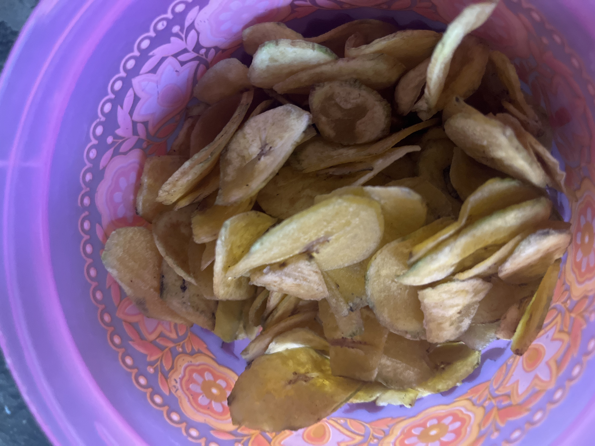 A close-up, top-down view captures a collection of golden-brown banana chips piled inside a transparent purple plastic container. The container features a decorative band with a floral pattern in shades of pink and orange around its upper edge. The banana chips are sliced thinly and appear to be crispy, with some individual chips showing slight browning or darker spots, indicative of frying. The lighting in the image is diffused, highlighting the texture and slight sheen of the chips. The scene is static and appears to be indoors.