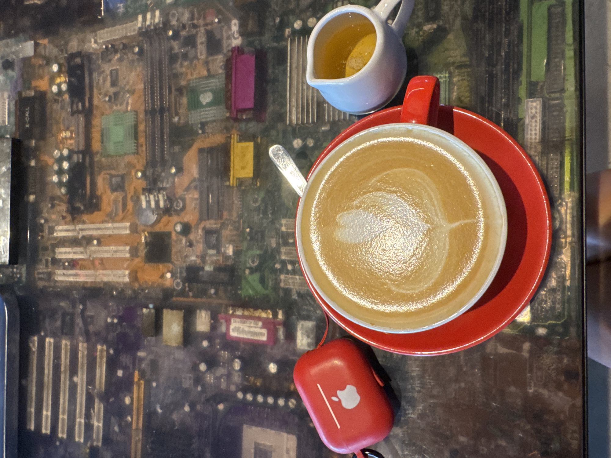 A red Apple AirPods Pro case is positioned to the left of a red saucer supporting a white cup filled with latte art. To the right of the cup, a small white pitcher holds a yellow liquid, possibly milk or creamer. The table's surface is a transparent pane displaying an assortment of computer motherboards. The image was captured at a coffee shop in Kuta Selatan, Indonesia.