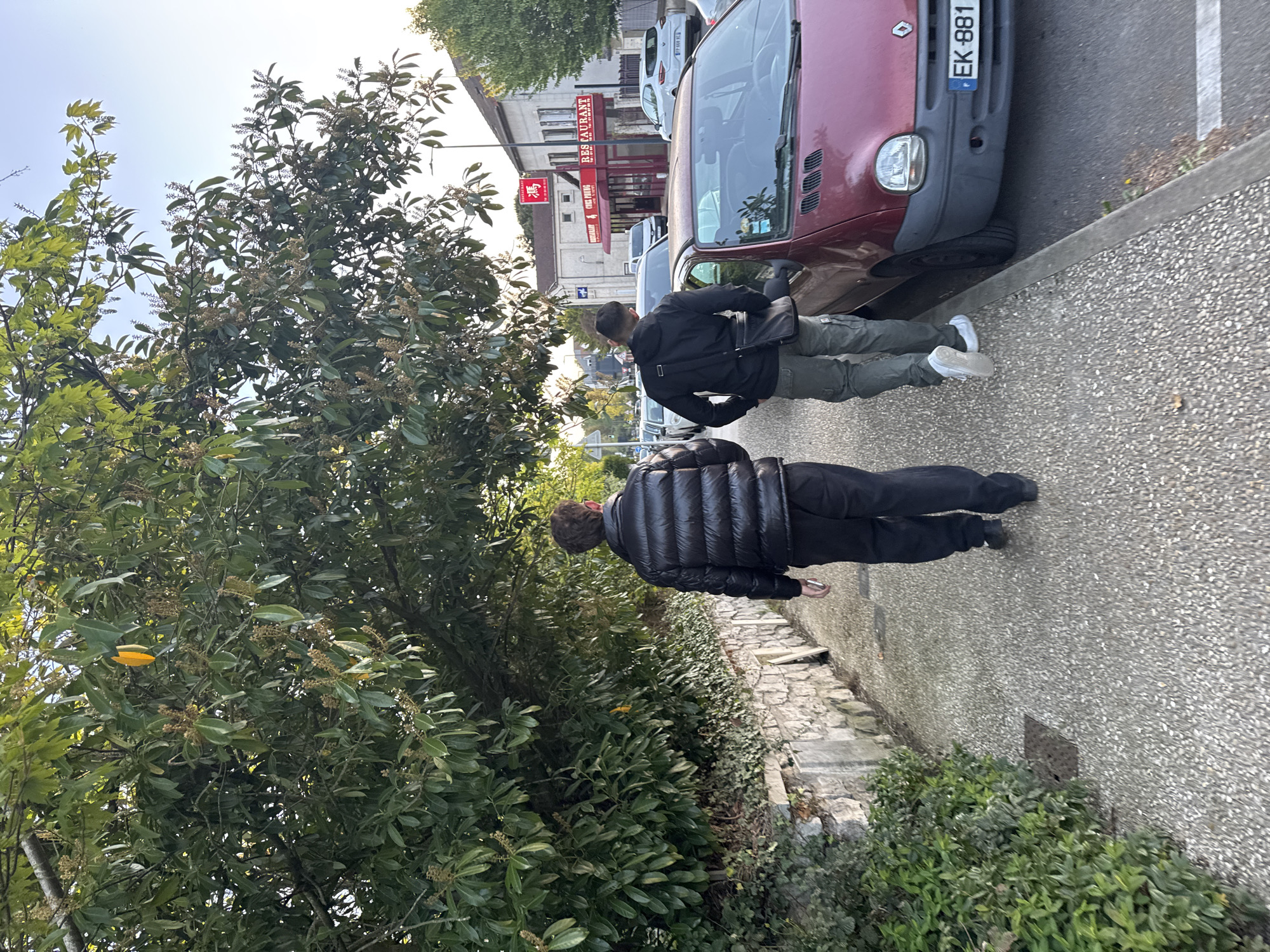Two individuals are captured walking along a paved sidewalk in Bures-sur-Yvette, France, on what appears to be a clear, bright day. The image is taken from behind the two subjects, looking down a street lined with parked cars and buildings.
The person in the foreground wears a black puffer jacket, black pants, and dark shoes. The second person, slightly ahead, is dressed in a black jacket, olive green or khaki cargo pants, and white sneakers. This individual also carries a black messenger bag slung over their shoulder.
To their right, a red car, identifiable as a Renault Twingo, is parked at the curb. Its French license plate, "EK-881," is clearly visible. Other cars are parked further down the street. The sidewalk is made of grey aggregate pavement, and a low stone wall separates it from a patch of green bushes.
In the background, a building features several signs, including "RES RESTAURANT" and "CHEF PENG" along with Chinese characters, indicating an Asian restaurant. The general setting suggests a commercial or residential street in a town or suburb. Lush green trees and bushes with small berries or flowers fill the left side of the frame, under a bright, clear sky.