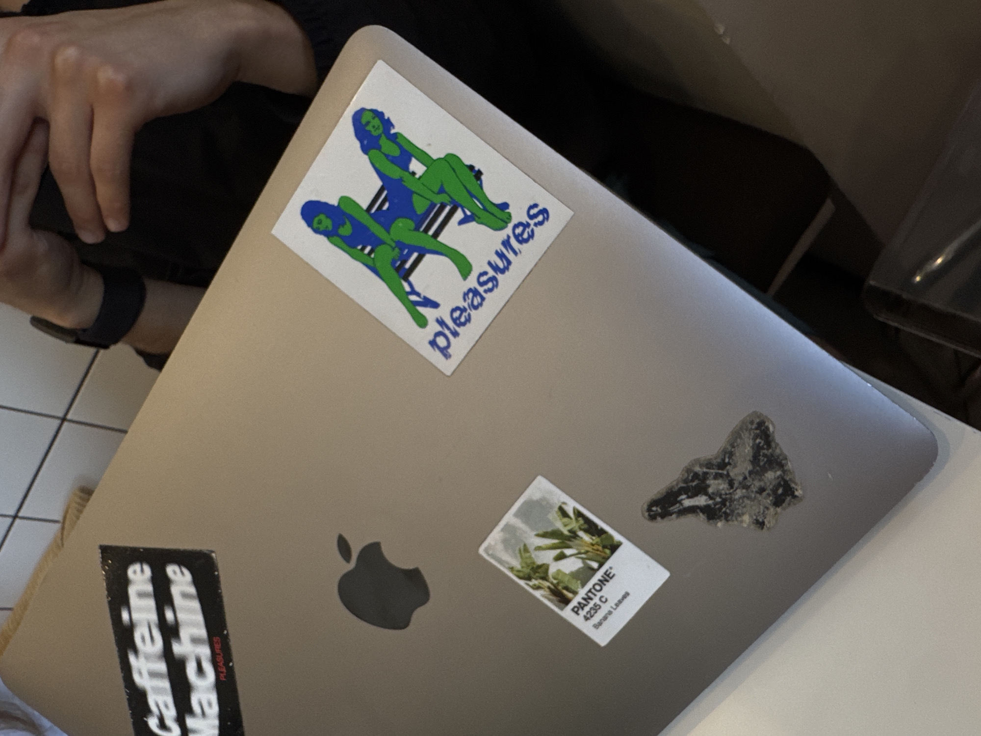 A slightly angled, close-up shot captures a silver laptop, likely a MacBook due to the visible Apple logo, resting on a light-colored surface, possibly a table or desk, in an indoor setting in Puebla, Mexico. On the left side of the frame, a person's hands and lower arm are visible, clad in a dark long-sleeved garment and a dark wristband. The background is dark and out of focus, suggesting ambient indoor lighting.
The laptop's lid is customized with several stickers. A prominent white rectangular sticker near the top features two stylized female figures with green bodies and blue swimsuits, reclining on striped chairs. Below them, the word "pleasures" is written in blue. Toward the bottom left, a black rectangular sticker displays "Caffeine Machine" in white text, with the word "PLEASURES" in smaller red letters beneath it. Another small white rectangular sticker shows an image of green leafy plants alongside the text "PANTONE® 4235 C Banana Leaves." An additional irregular-shaped, dark, textured sticker is positioned near the Apple logo.
The scene portrays a personal computing setup, suggesting activities such as work, study, or leisure. The visible hand indicates human presence and potential interaction with the device. Details like the white tiled floor with dark grout lines are partially visible in the far left. The array of stickers offers insights into the owner's interests, encompassing design, color, and potentially coffee culture. The image provides no specific time of day, weather, or emotional cues.