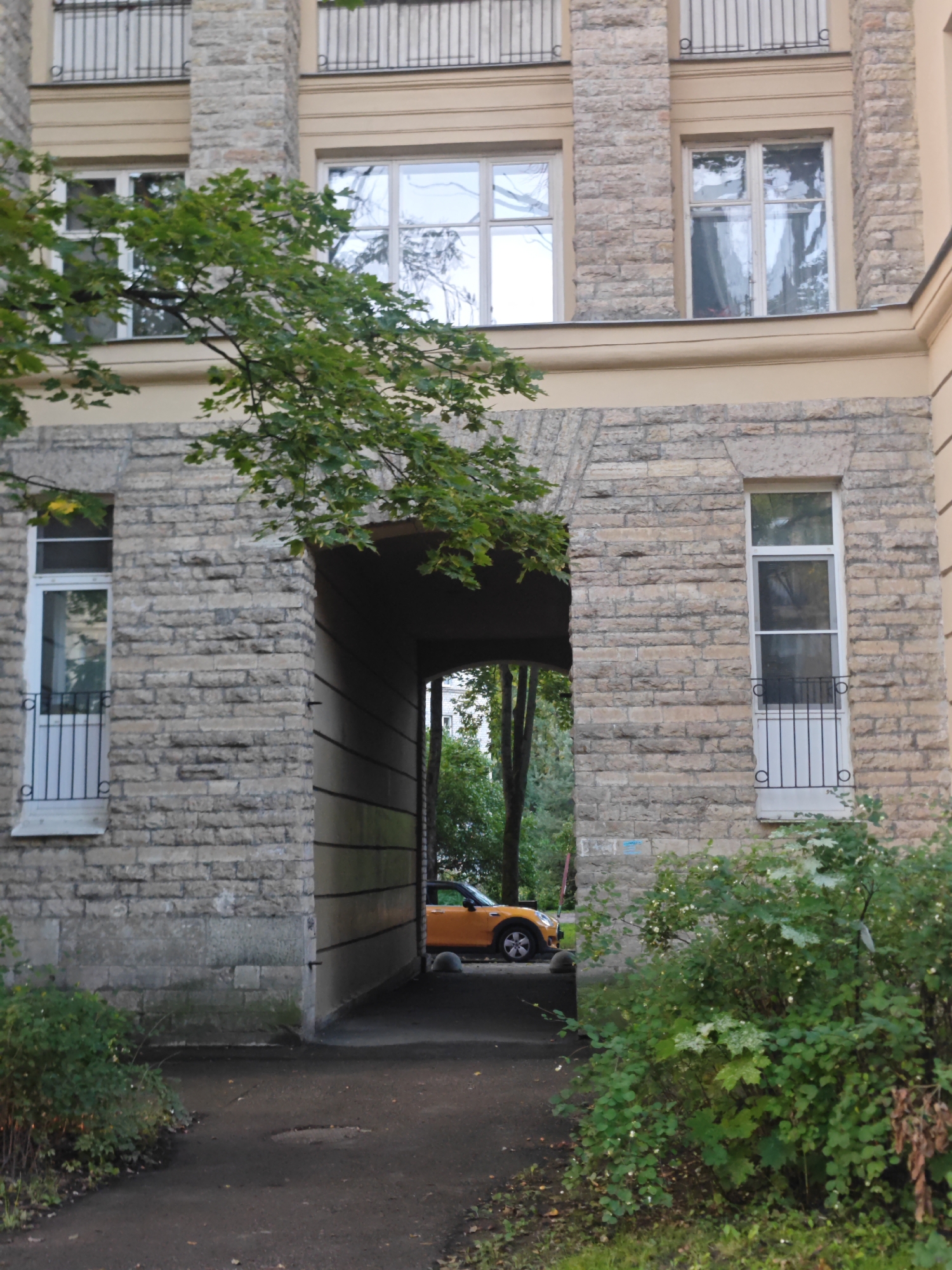The image depicts an exterior view of a building in Pushkin, Russia, likely a residential or administrative structure. The building is constructed with stone blocks, featuring multiple windows and a distinct archway or passage. Green foliage from trees and bushes is visible in the foreground and around the archway. Through the archway, a glimpse of an orange car parked on a paved surface is visible. The scene appears to be outdoors, with natural light illuminating the area, suggesting it was taken during the daytime. No people are present in the frame. The overall impression is of an urban architectural setting with natural elements.