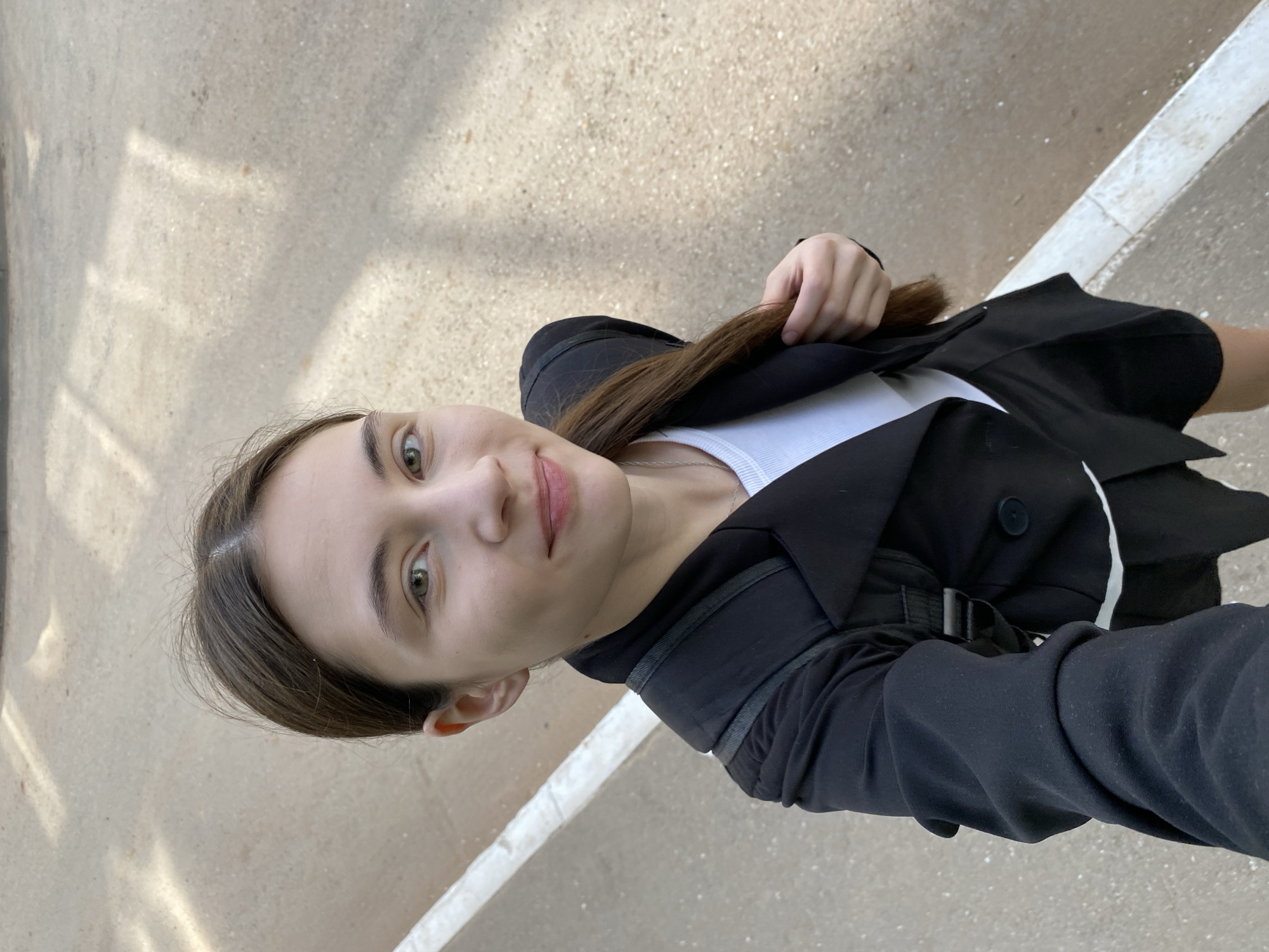 A young woman stands outdoors in Votkinsk, Russia. She wears a white shirt, a black blazer, and a black backpack. She is holding a strand of her brown hair. The ground is light-colored concrete with a white painted line.