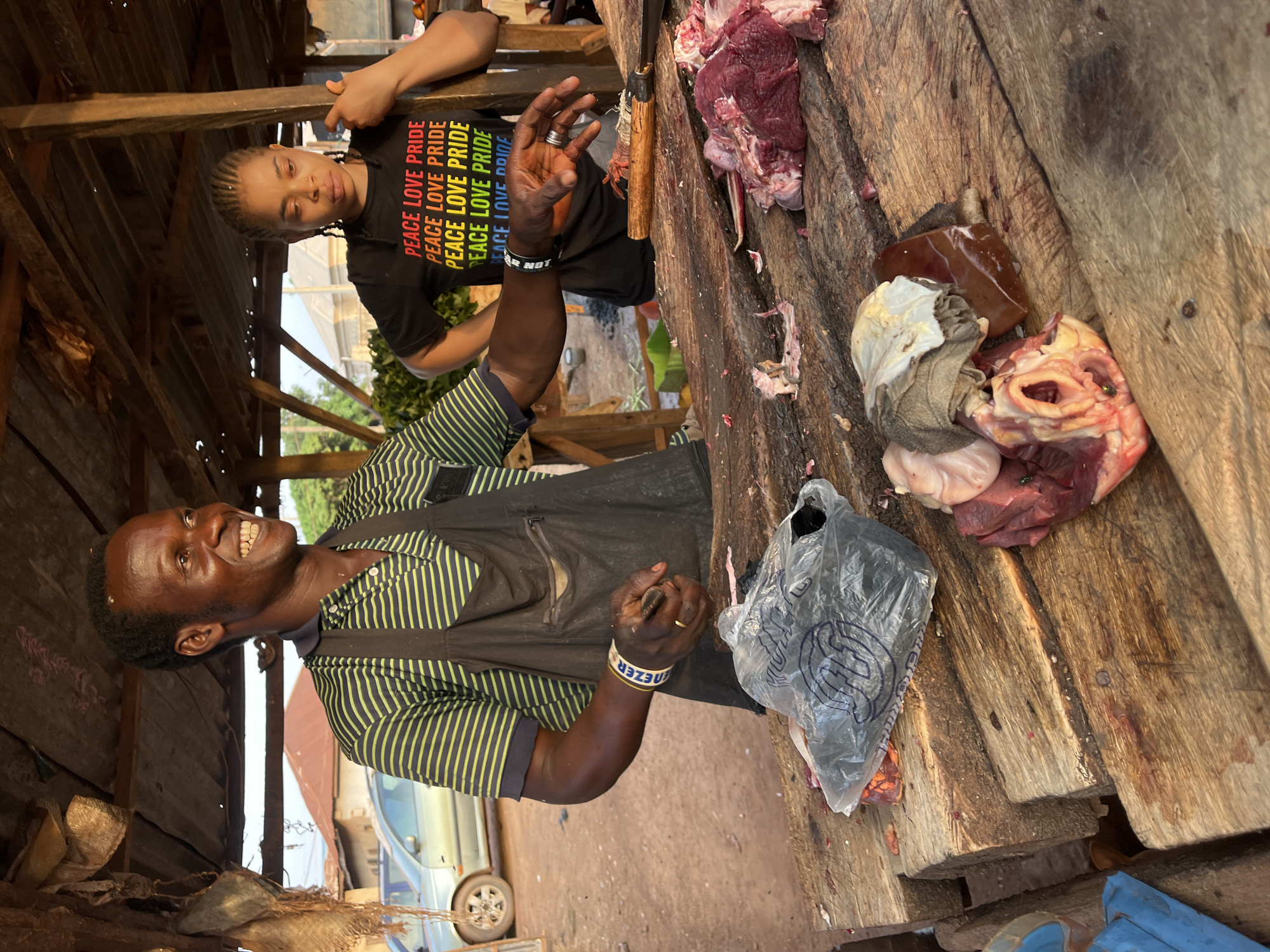The photograph captures a butcher, smiling broadly, standing behind a rough wooden table loaded with various cuts of meat, including what appears to be a liver and a heart, in a rustic setting that suggests a meat market. The scene is located in Abuja, Nigeria. The butcher is clad in a striped shirt and a black apron, wearing a wristband. Next to him, a plastic bag sits on the table among the meat. In the background, a second person is seen, wearing a t-shirt with "PEACE LOVE PRIDE" printed on it multiple times. Behind the butcher is a simple structure made of wood and other materials. A car is partly visible in the background.