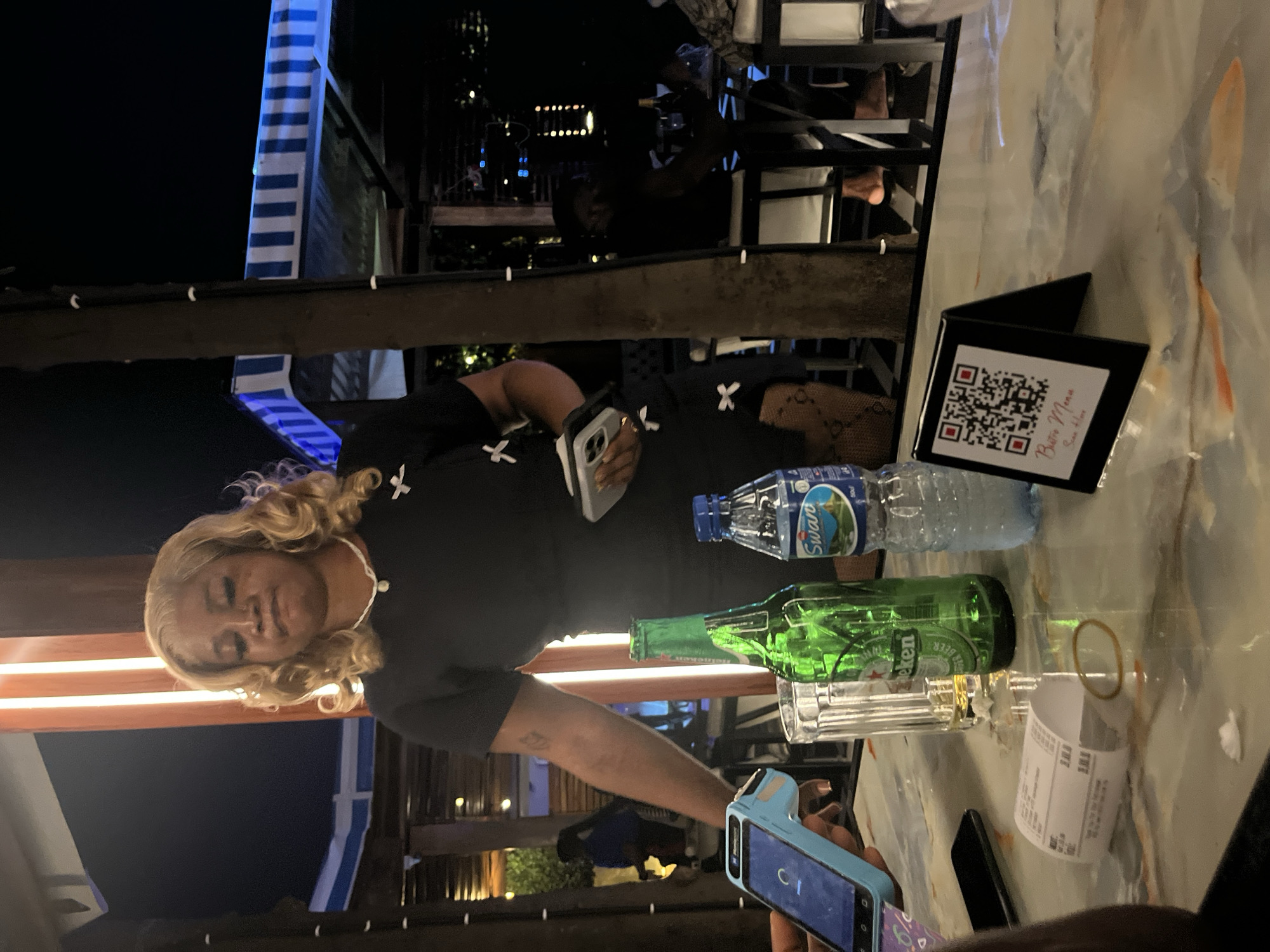 A woman with blonde, curly hair and fair skin is standing in what appears to be an outdoor dining establishment in Abuja, Nigeria. She is wearing a black dress with white cross embellishments, a white beaded necklace, and fishnet stockings. She is holding a smartphone in her right hand and a small device, possibly a credit card reader, in her left. In front of her on a marble-topped table are a green beer bottle, a bottle of Swan water, and a glass. A receipt is also present on the table. A QR code menu is displayed on a small black stand in the foreground. The background shows other patrons and lighting that suggests it is nighttime.