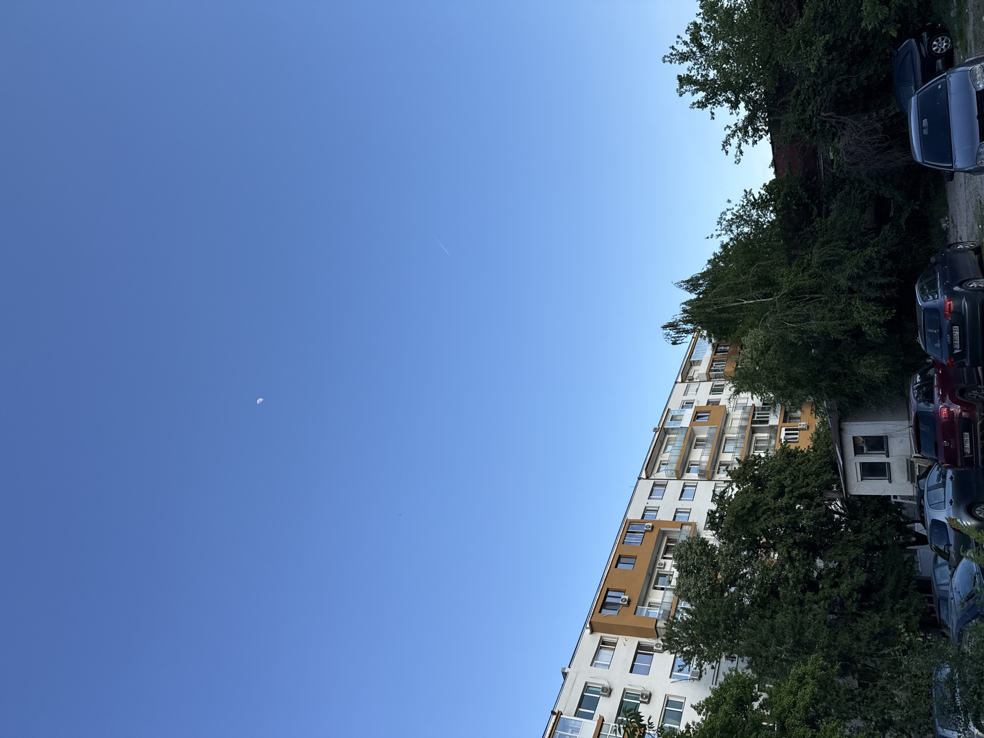 The image shows a partly cloudy sky in Lovech, Bulgaria, with a crescent moon visible in the upper left. A contrail from a plane is also present. In the lower right, a multi-story apartment building is visible, partially obscured by trees. Cars are parked next to the building. The building has a white facade with brown accents around some windows and balconies. Air conditioning units are attached to several of the windows.