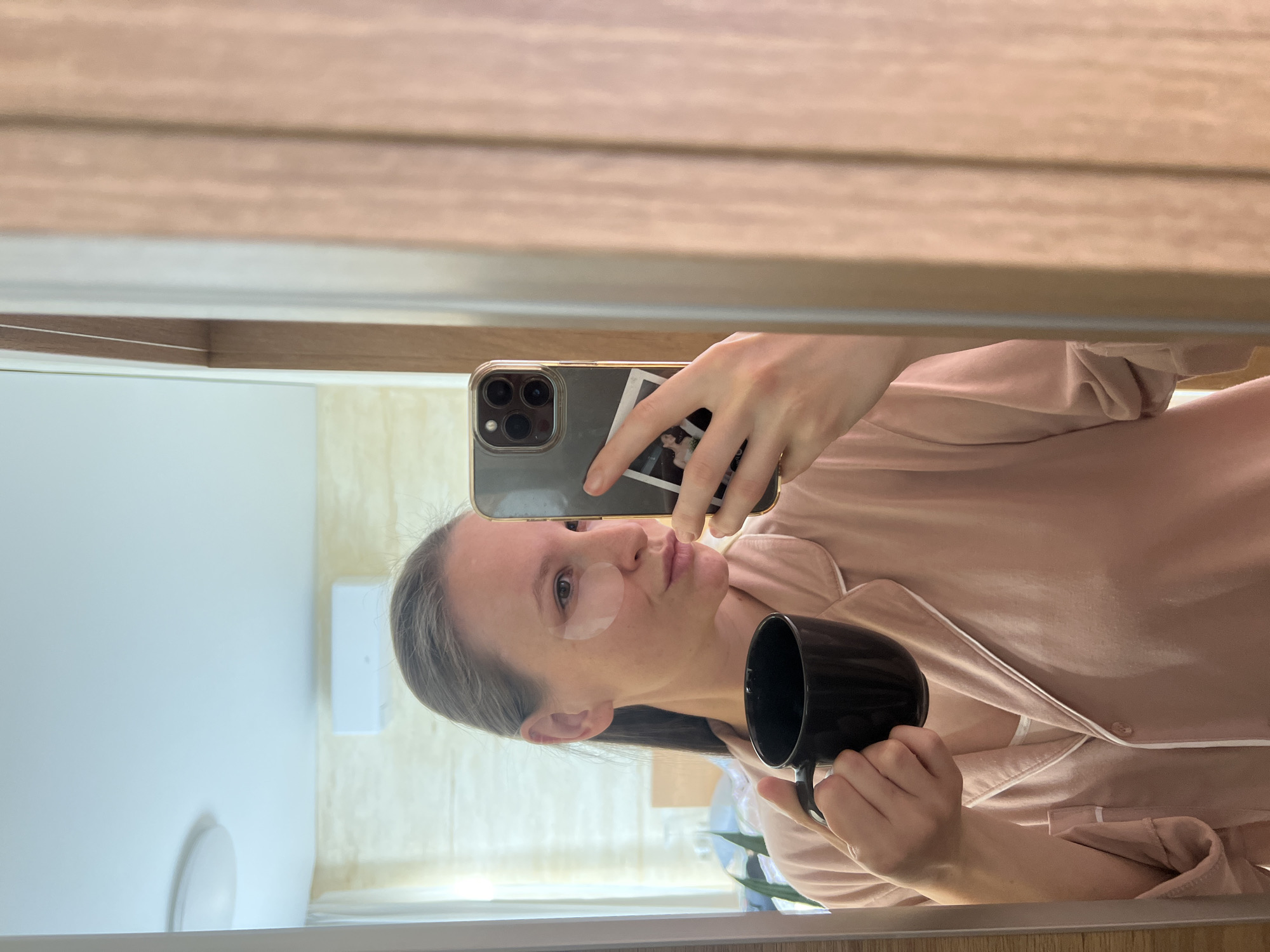 A woman is taking a selfie in a mirror. She is wearing a pale pink pajama top with white trim and has a round eye patch under her left eye. She is holding a black mug in her right hand and a smartphone in her left hand. The phone has a small picture attached to the back. The mirror is framed in wood, and the background reflection shows a white wall, a white light fixture, and a beige wall with vertical lines. There is no indication that this image was taken in Gelendzhik, Russia.