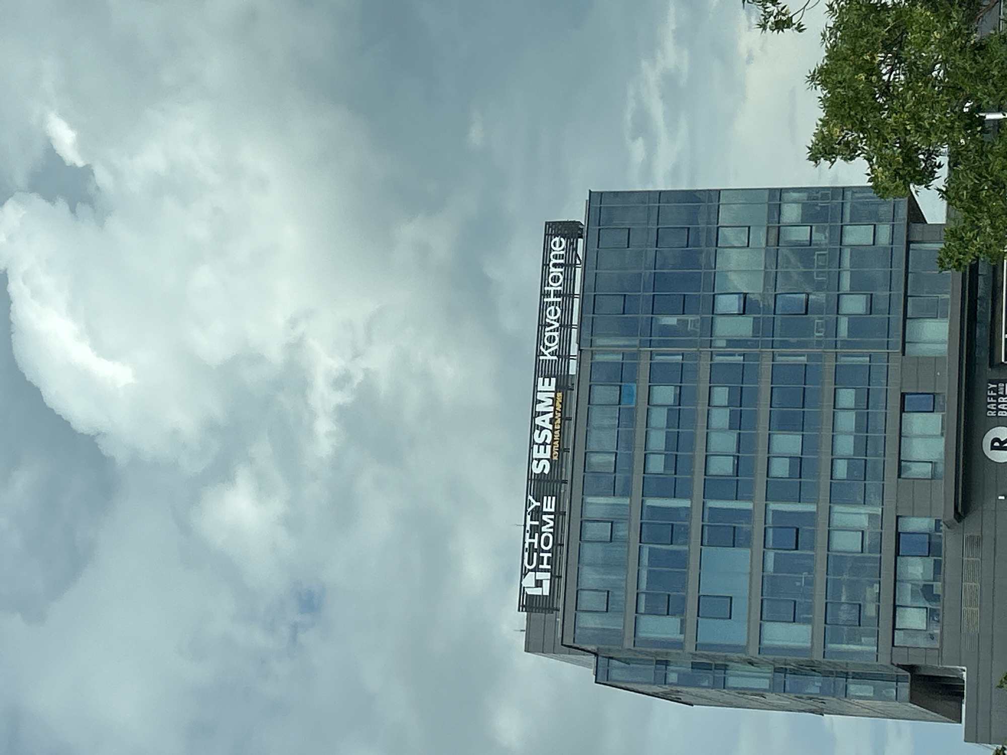 The image shows a modern glass-fronted building in Sofia, Bulgaria, under a cloudy sky. The building has signage at the top that reads "CITY HOME SESAME Kave Home" with a smaller line of text below "SESAME" that appears to be in Cyrillic script. The lower part of the building has a darker facade with visible windows. A green tree is visible in the lower right corner of the image. The overall scene is a cityscape view.
