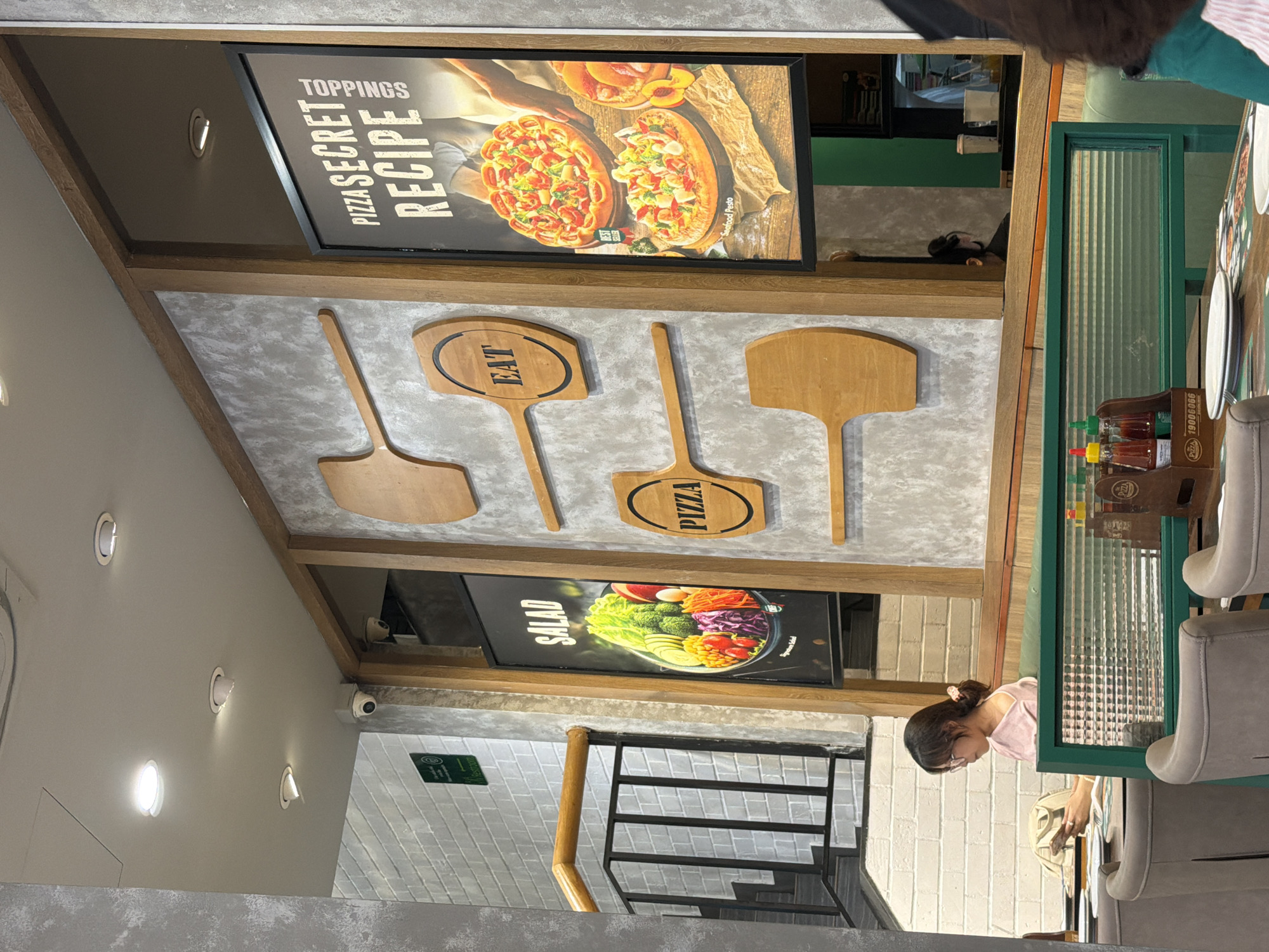 Captured indoors, the image shows an establishment likely in Đà Nẵng, Vietnam, given the style. An unidentified female, possibly a patron or employee, is present. The interior décor includes pizza and salad advertisements alongside wooden pizza paddles bearing "Eat" and "Pizza" engravings. Furniture is visible, specifically chairs. The establishment has staircase leading up. Recessed lighting is installed throughout the building.