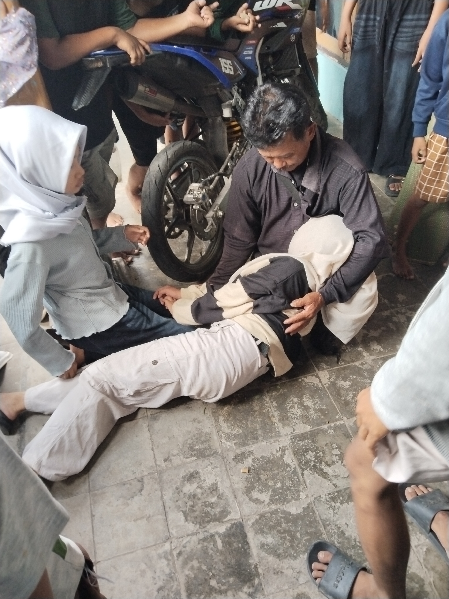 The image depicts a scene in Mojodoyong, Indonesia, where a person lies prone on a tiled floor, receiving attention from another person kneeling beside them. The person on the floor wears light-colored pants and a top with black and beige sections, and a hijab covering their head. The kneeling person, dressed in a dark button-down shirt, is touching the person on the floor with their hands. A person wearing a white hijab and a light blue shirt is also seated next to the person on the ground, holding their hand. A motorcycle is partially visible in the background, along with several other people standing around, observing the situation. Some of them are wearing casual clothing like shorts and sandals. The environment appears to be an outdoor space, with the tiled floor as the ground surface.