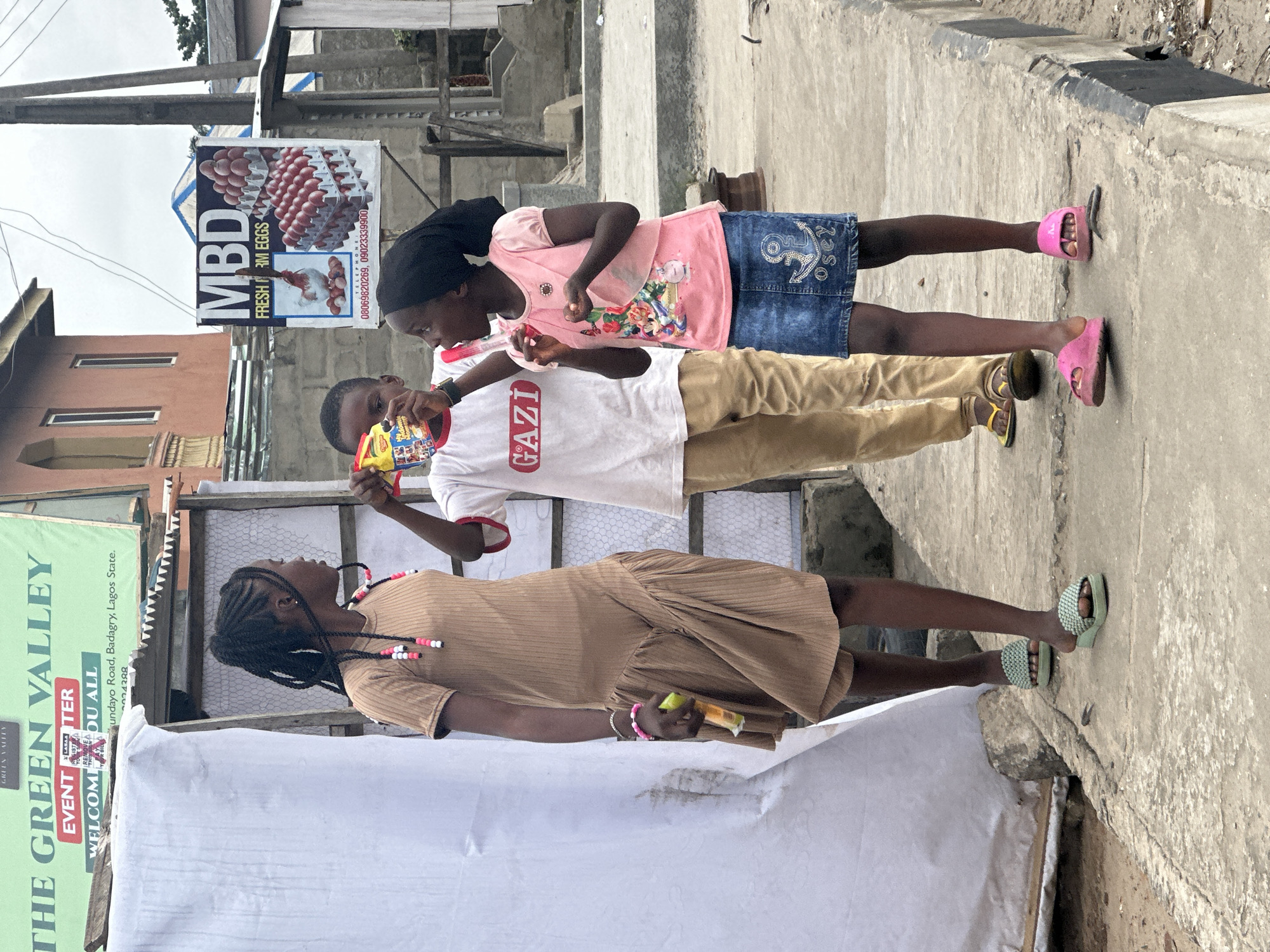 The image shows three children standing on a concrete pathway in Badagry, Nigeria. A young girl on the left, wearing a tan ribbed dress and green sandals, holds a yellow and red packaged item in her right hand. She has braided hair adorned with pink and white beads. In the center, a boy wearing a white t-shirt with "GAZI" printed on it and khaki pants holds up a yellow package. To the right, a girl in a pink t-shirt and a denim skirt, with a black headscarf, holds a pink object. She is wearing pink flip-flops. Behind the children, a large green banner with "THE GREEN VALLEY EVENT CENTER" is visible, along with a sign advertising "MBD FRESH FARM EGGS" with images of eggs and a chicken. The environment appears to be an urban setting with buildings and a street curb.