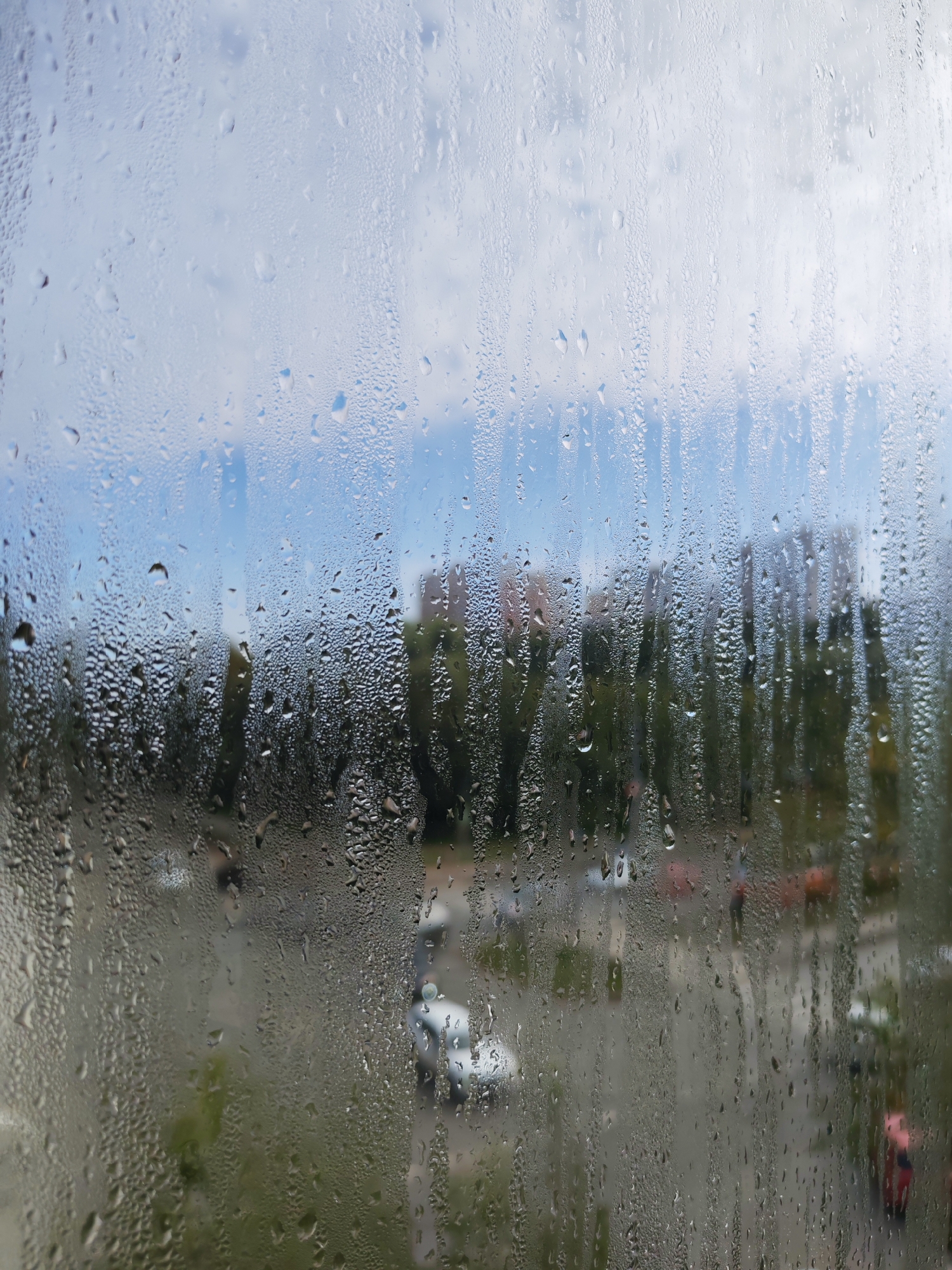 This is a photograph of a window covered in condensation, looking out onto a city scene. The condensation obscures much of the detail of the exterior view. In the background, there appear to be several multi-story buildings, some with reddish-brown facades, surrounded by trees with green foliage. A paved area, likely a street or parking lot, is visible below the trees. Several vehicles, including a white car and potentially others in red and orange, are present in the scene. The sky above is cloudy and appears to be a pale blue. The image was taken in Izhevsk, Russia, likely from a residential or office window, suggesting a view of an urban or suburban area within the city. The condensation on the glass creates streaks and droplets, giving the impression of a rainy or humid day.