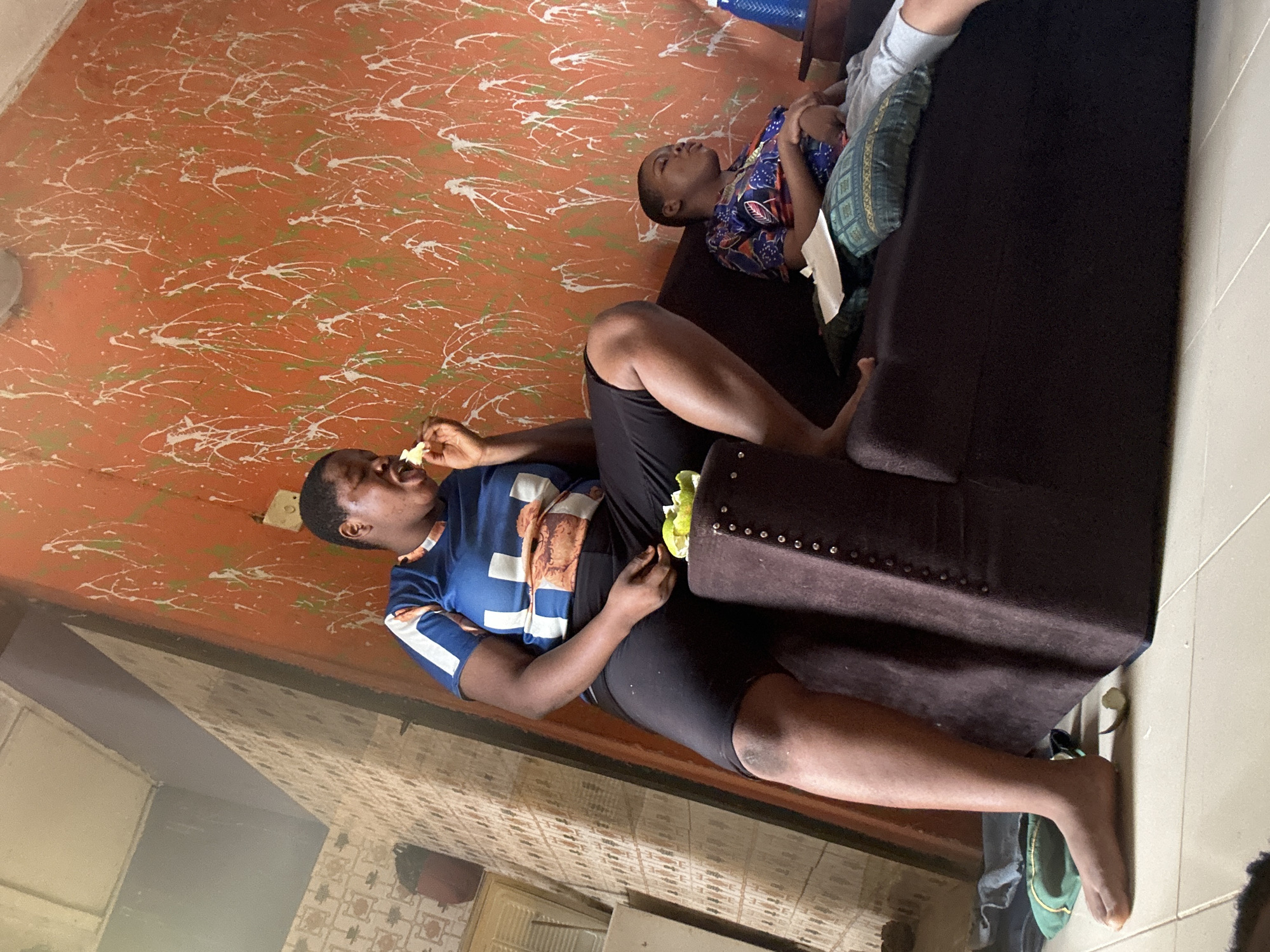 The image shows two individuals inside an unknown location, Unknown, Unknown. One person is lying on a dark brown sofa, possibly asleep or resting, wearing a blue patterned shirt and green patterned shorts. Another person is sitting on the sofa, eating something, wearing a blue and white patterned shirt and black shorts. The room has a distinctive orange wall with white and green splatter paint design. The overall lighting suggests a daytime scene.