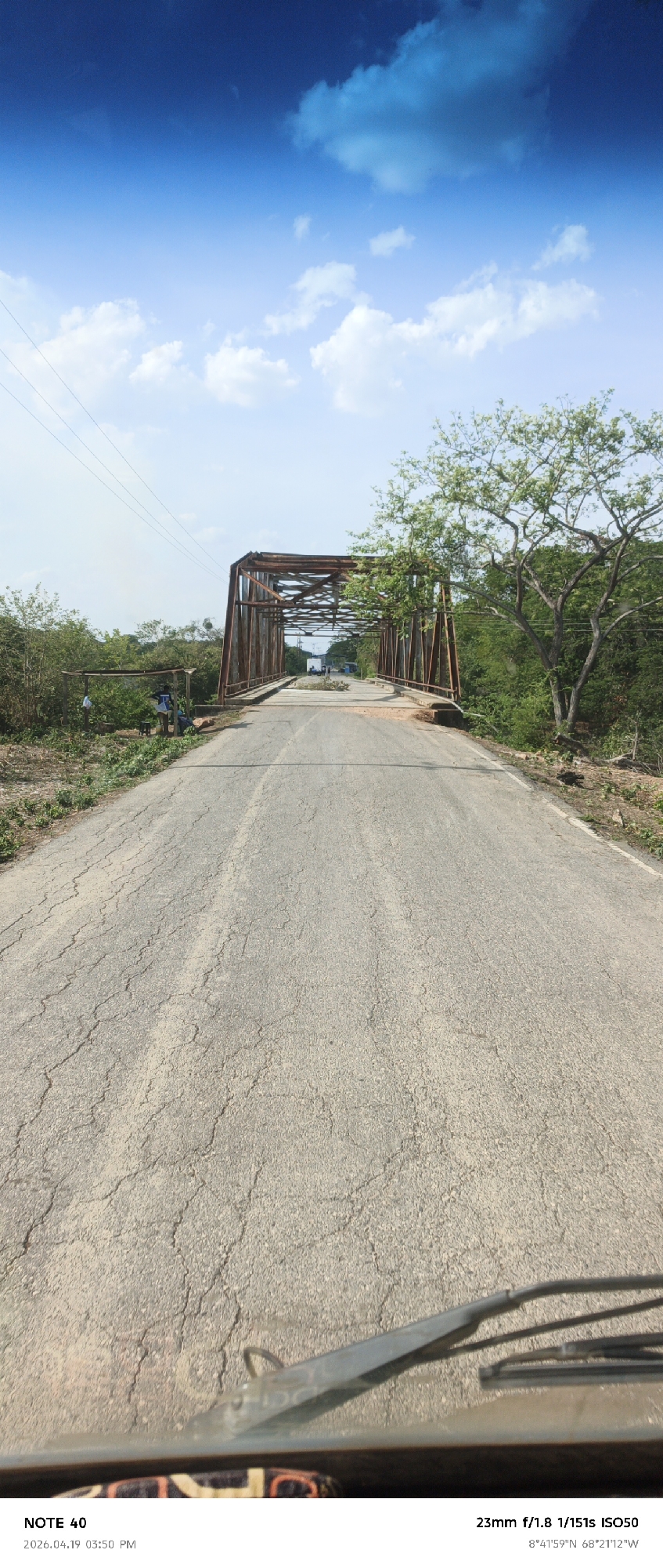 The image shows a bridge in the rural area of El Baúl, Venezuela, taken from a vehicle traveling on a cracked asphalt road. The bridge has a rusted metal frame, and vegetation surrounds it. Two individuals are near the left side of the road close to the bridge entrance. The sky is blue with scattered clouds. A vehicle windshield and wipers are visible at the bottom of the image.
