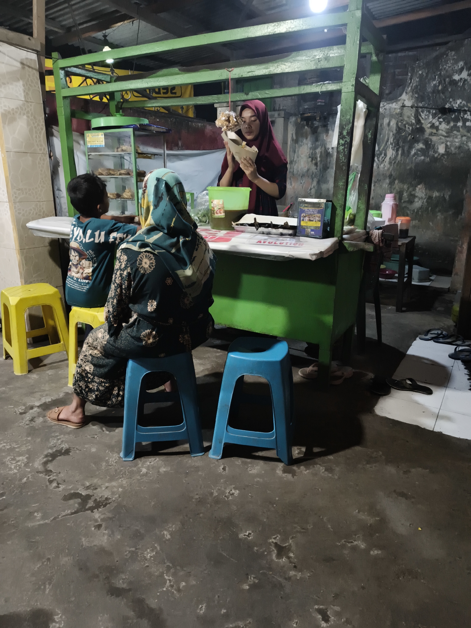 The image depicts an outdoor food stall scene in Sidorejo, Indonesia. A woman wearing a red hijab and a dark top stands behind a green counter, holding a food item in a paper cone and appearing to serve it. To her left, a young boy in a teal t-shirt sits on a yellow stool facing the counter. In the foreground, a woman wearing a teal and patterned hijab and matching patterned pants sits on a blue stool, facing away from the camera. The stall has a green frame and a white countertop, with food displayed in a glass cabinet behind the woman. The ground is concrete, wet in patches, and littered with several pairs of flip-flops. The overall environment suggests a casual, street-level food vendor operation.