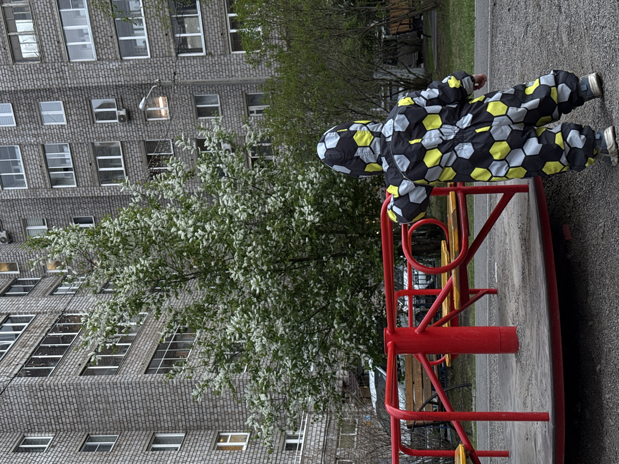A child wearing a black, yellow, and white soccer-ball-patterned jumpsuit is playing on a red and yellow playground merry-go-round in what appears to be a courtyard in Krasnoyarsk, Russia. In the background is a large, Soviet-era, brick apartment building, as well as blossoming trees and other greenery. The scene is slightly rotated clockwise. The ground is paved with a dark gray material.