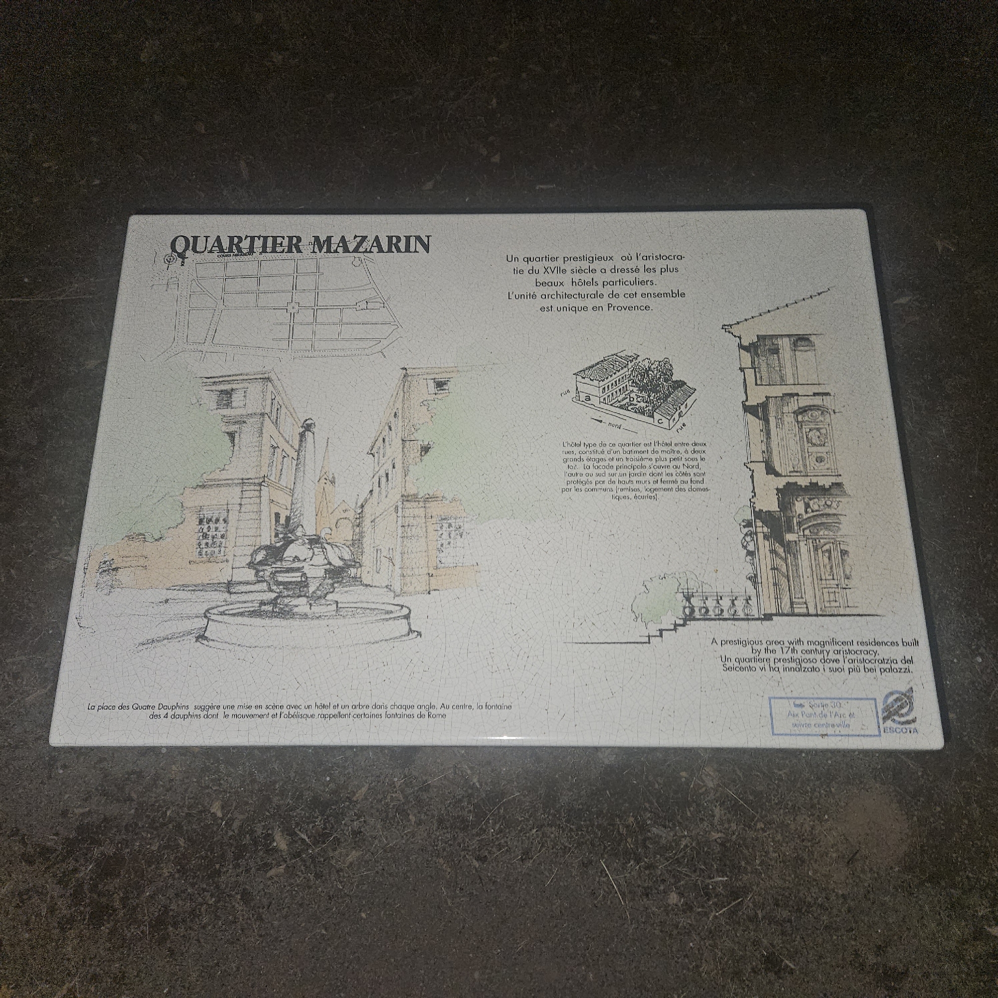 The image shows a white ceramic tile, likely a plaque or sign, set against a dark, textured surface. The tile features a detailed ink drawing and some colored accents depicting an urban scene with architectural elements. The title "QUARTIER MAZARIN" is prominently displayed in black lettering at the top left.
To the right of the title, a French text describes the Mazarin Quarter as a prestigious area where the 17th-century aristocracy built magnificent private residences, noting its unique architectural unity in Provence. Below this, an aerial view sketch illustrates a street layout with houses and a central courtyard.
The main part of the tile depicts a street scene, possibly a square, with a fountain in the center. Buildings with multiple stories and windows are drawn around the square. A sketch of a detailed building facade with ornate architectural features is shown on the right side of the tile. Color is used sparingly, with some areas of light green and a peachy hue on the buildings, and darker tones for the fountain and some architectural details.
At the bottom of the tile, English and Italian text reiterate the description of the prestigious area with magnificent residences built by the 17th-century aristocracy. A small blue box at the bottom right contains text that appears to be a route or address designation, reading "Sortie 30" and "Aix Pont de l'Arc et suivre centre ville". This information, combined with the reference to Provence in the text, suggests the location is in or near Aix-en-Provence, France. The presence of the "ESCOTA" logo indicates this is likely related to a regional road management authority.
The overall scene is a descriptive informational plaque, likely for tourists, detailing a specific historical district. The background suggests the plaque is placed outdoors or on a surface that resembles cobblestones or rough ground.