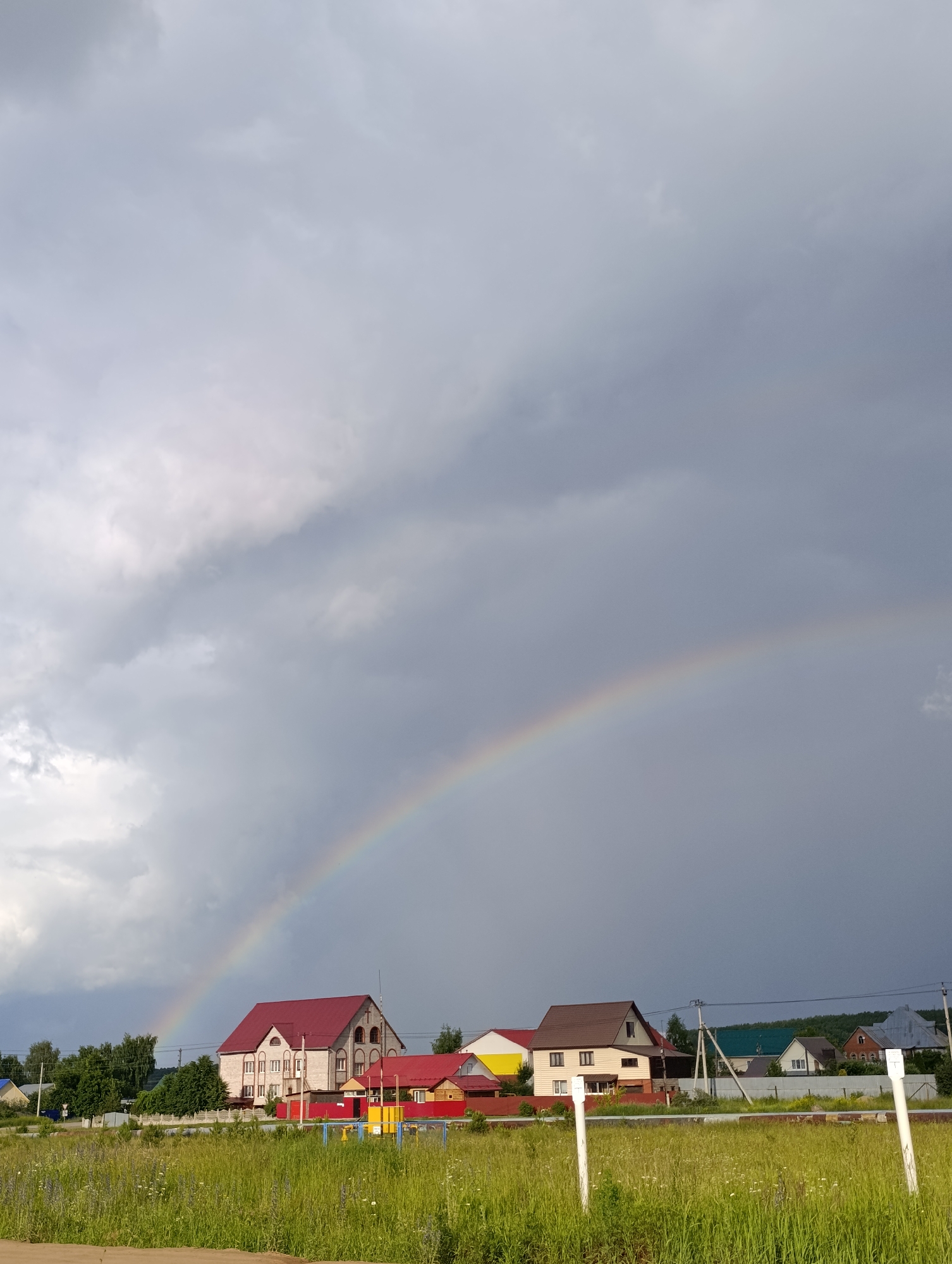This is a photograph of a rural landscape with a rainbow in a cloudy sky. In the foreground, a grassy field with wildflowers is visible. Behind the field, there is a row of houses with red and brown roofs. The houses appear to be residential properties. To the right of the houses, there is a playground with yellow and blue structures. A white fence runs along the edge of the field. The sky is dominated by dark, heavy clouds, with a bright rainbow arcing across it. The overall impression is of a recent rainfall followed by a break in the clouds. The location is the city of Chaikovsky, Russia. There are no people visible in the image.