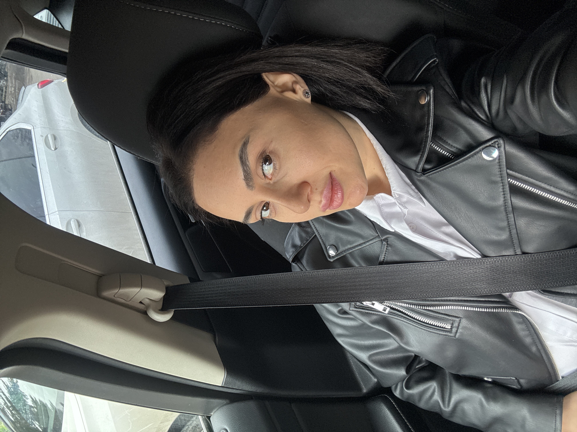 A woman with dark hair, wearing a black leather jacket over a white shirt and a seatbelt, sits inside a car. The car is parked on a street in Tbilisi, Georgia. A white car is visible through the window.