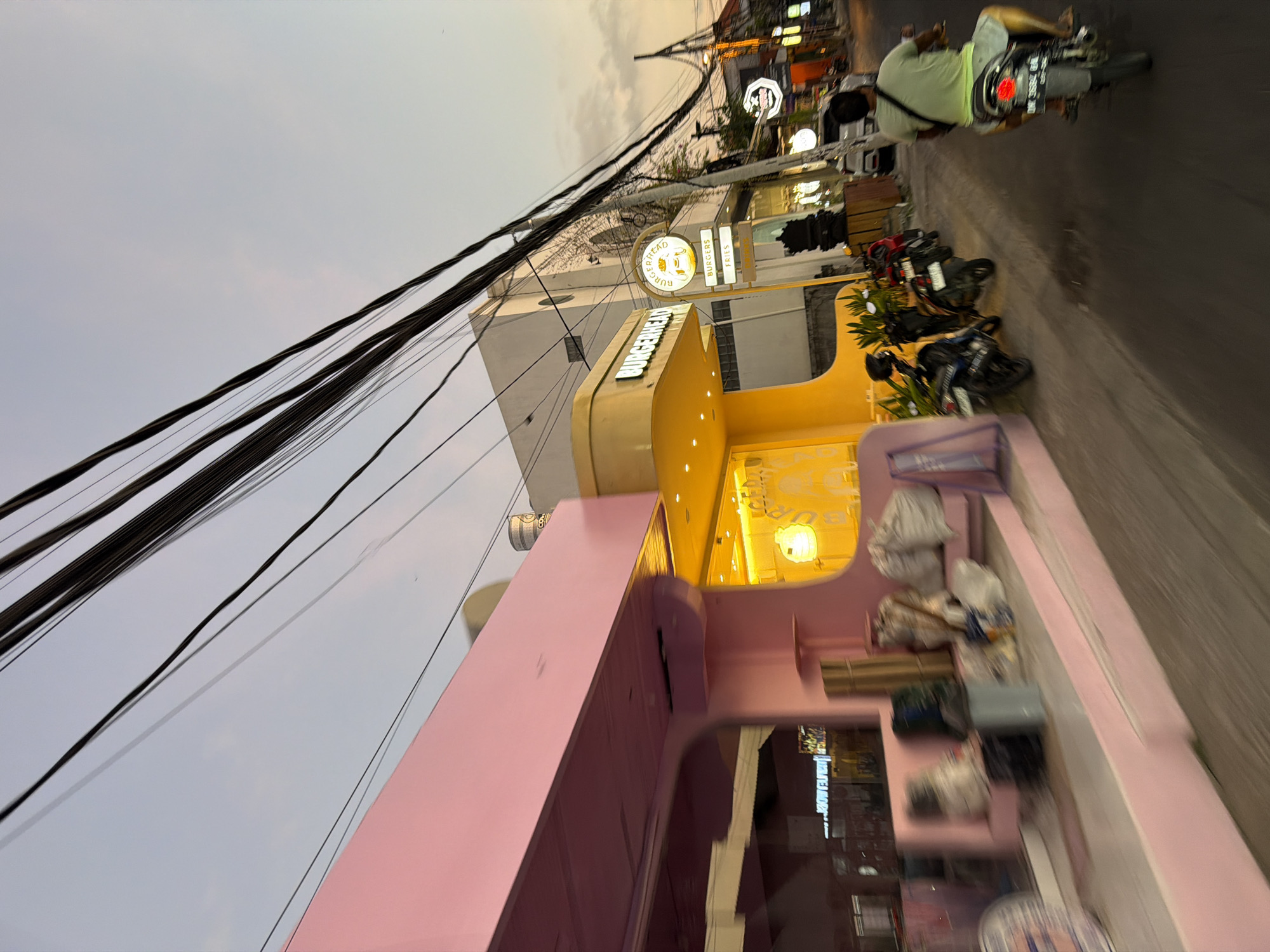 The image depicts a street scene at dusk in North Kuta, Bali, Indonesia, specifically showing the area of Burgerhead Bali on Gg. Janji, Canggu. The sky above transitions from light blue to a pale orange-pink at the horizon. Numerous black power and communication lines are strung diagonally across the upper portion of the frame.
On the left, a two-story building features an entirely pink exterior, with large windows reflecting ambient light and indistinct interior activity. Its base includes a stepped platform where multiple objects, including bundles wrapped in white and brown material, a blue container, and various bags, are stacked. A cylindrical metallic object is visible on the edge of the pink building's roof.
Adjacent to the pink building on the right, a bright yellow structure with a rounded, modern design houses "BURGERHEAD." The restaurant's name is displayed in white text on the yellow facade. In front of this building, two illuminated "BURGERHEAD" signs are visible: a circular sign featuring a burger graphic and text, and a rectangular sign below it stating "BURGERS" and "FRIES." Green foliage is planted at the base of the yellow building. Several motorcycles are parked on the sidewalk in front of the yellow building.
Further down the street, a white building with an arched window and some vine growth is visible. The road surface is asphalt. A person riding a motorcycle, wearing a light green t-shirt and light-colored shorts, is moving away from the viewer on the right side of the road. The motorcycle has a red taillight and a license plate partially visible as "DM 3684 BA." Other buildings and ambient streetlights are faintly visible further down the street on the right.