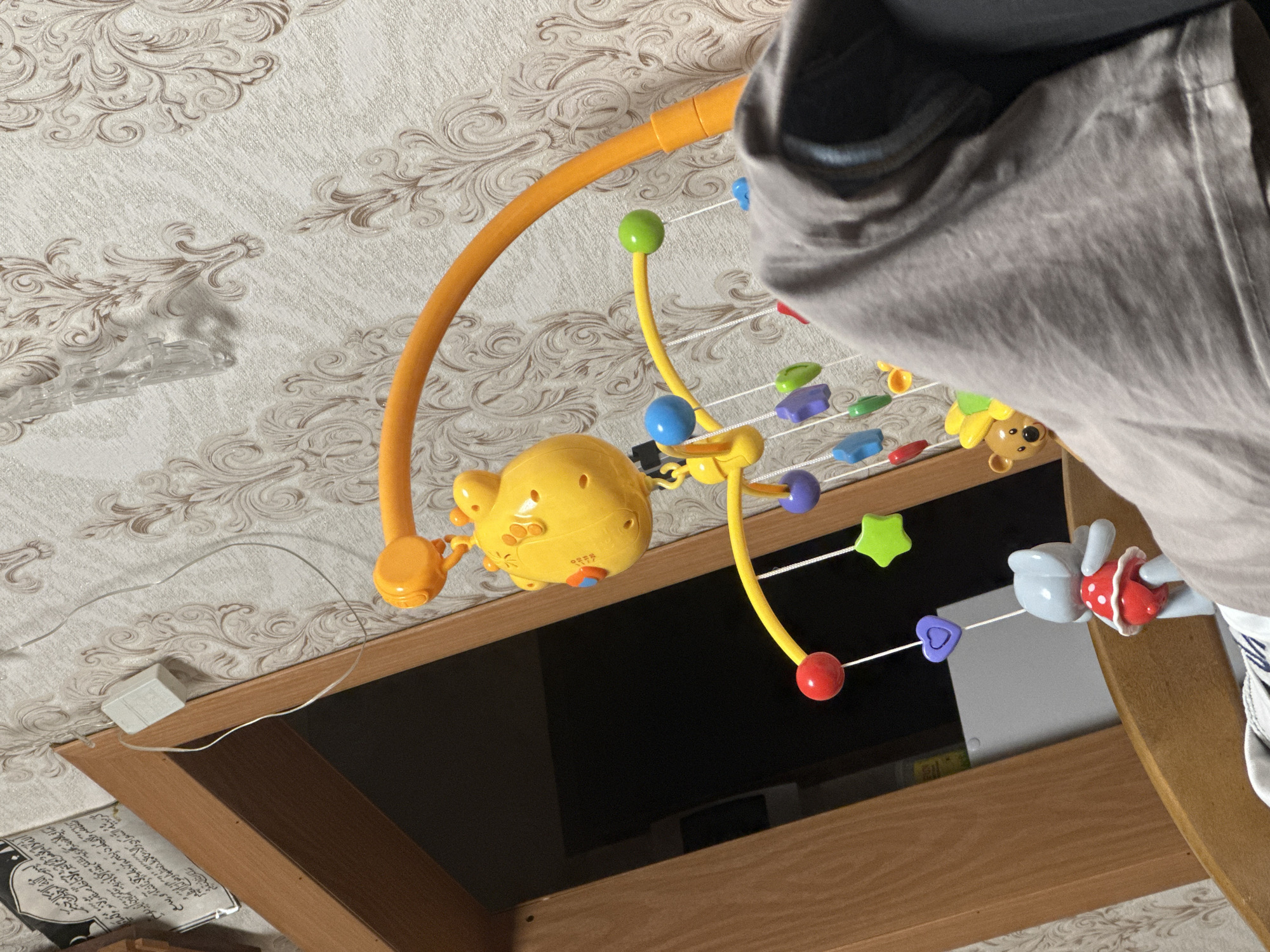 The image shows a close-up of a baby crib mobile, predominantly yellow and orange, with various hanging toys including a yellow giraffe head, colored balls, stars, and hearts. A small grey teddy bear and a grey mouse toy wearing a red polka-dot outfit are also visible. The mobile is attached to an orange plastic arm extending over the crib. In the background, there is patterned wallpaper and a wooden door frame. A piece of grey fabric is draped in the foreground, obscuring a portion of the crib. A piece of Islamic calligraphy is hanging on the wall to the left of the door frame. The scene appears to be indoors, likely a nursery. There is no indication of the city of Загородный, Russia.