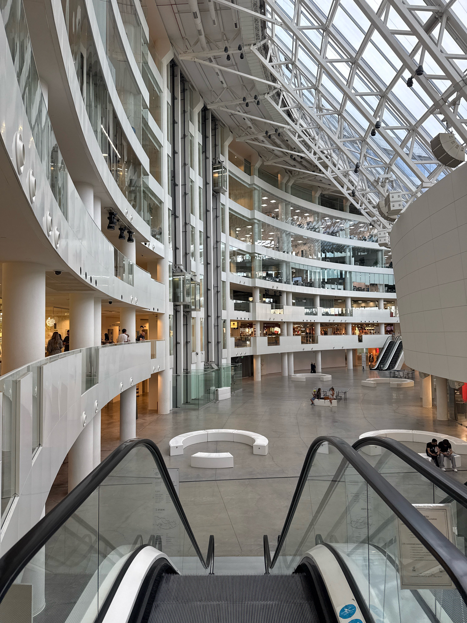 This image captures a wide-angle view of the interior of the Yeltsin Center in Yekaterinburg, Russia. The architecture features a striking modern design with a multi-level, open-plan atrium. Dominating the foreground are two escalators, with their glass railings reflecting the surrounding structure. The building's interior is characterized by curved white balconies, large glass windows, and a distinctive geodesic dome-like glass ceiling, allowing natural light to flood the space.
In the atrium, people are visible, some walking, others seated on modern white benches, and a few conversing. On the different levels, offices and retail spaces with glass facades are apparent. An elevator shaft is also visible, integrated into the building's vertical design. The overall impression is one of a spacious, contemporary public space, likely designed for cultural or commercial use. The environment appears clean and well-maintained, with polished concrete floors and a minimalist aesthetic.
