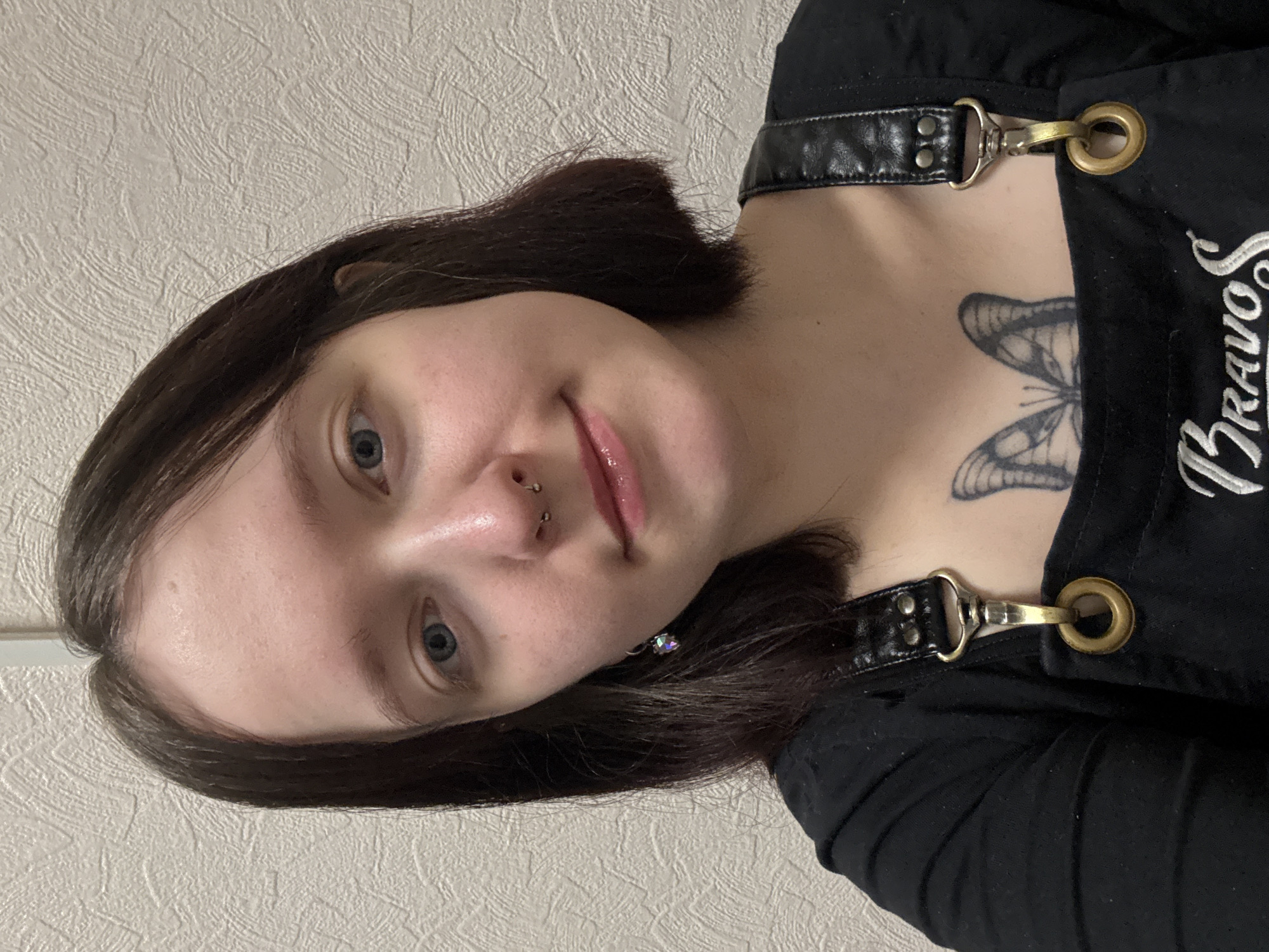 A rotated close-up portrait features a young person with short, dark brown hair and light blue eyes, gazing directly at the camera with a neutral to slightly smiling expression. The individual has a nose piercing and an earring in their right ear. They are wearing a black garment, possibly overalls or a pinafore dress, which has black faux leather straps secured with metallic clasps and brass-colored eyelets. A tattoo of a butterfly is visible on their upper chest/collarbone area. Partially visible white text, beginning with "Bravo" and ending with a stylized "S," is embroidered or printed on the black clothing. The background is a light-colored, textured wall, suggestive of an indoor setting. The overall presentation is a selfie taken at an angle, with the person's head tilted to the viewer's right. The context provided indicates the location is Chaikovsky, Russia.