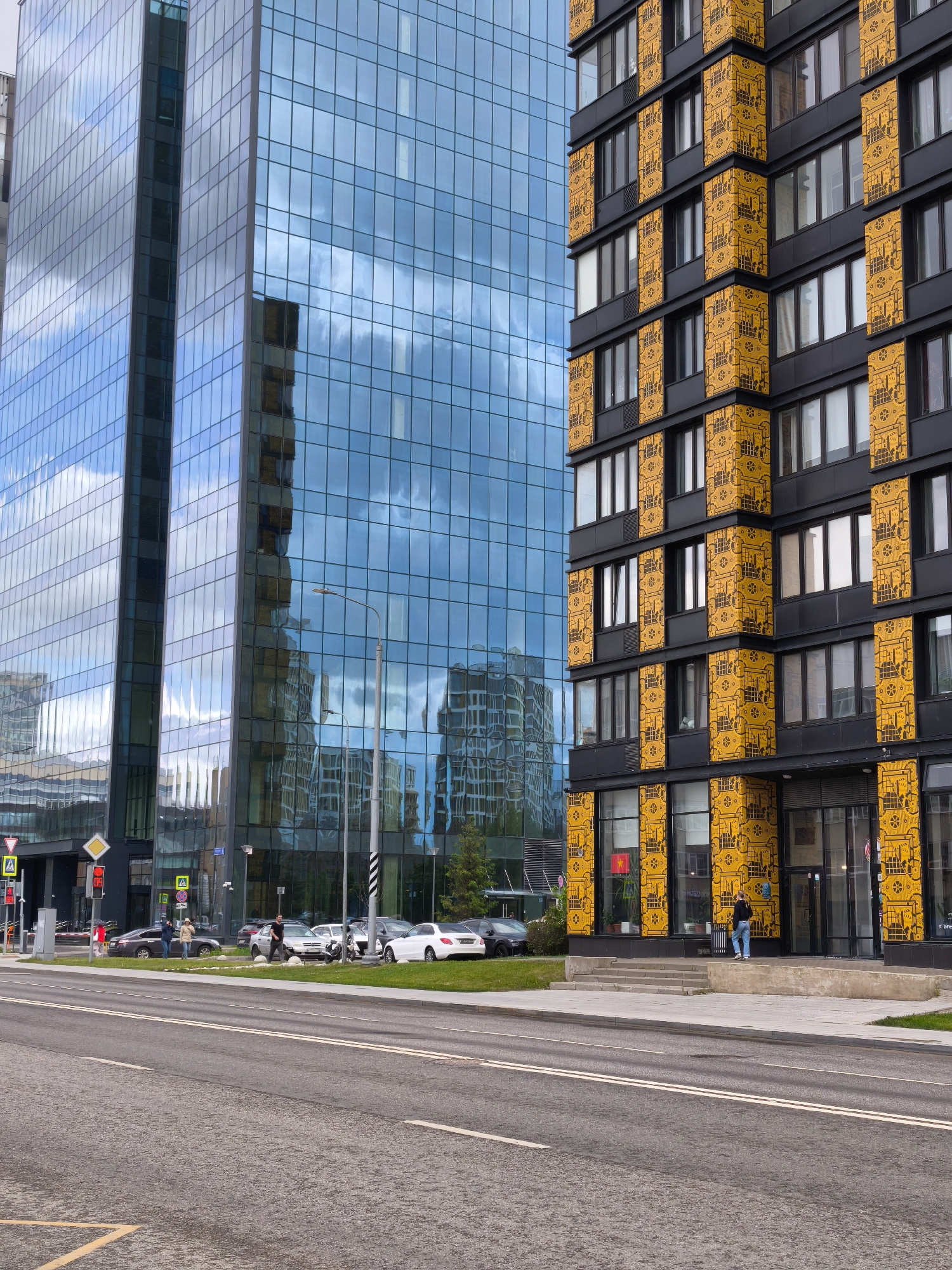 The image shows an urban scene in Moscow, Russia, featuring two prominent buildings. The building on the left is a modern glass skyscraper reflecting the sky and clouds. To the right stands a building with a dark facade adorned with yellow decorative panels.
A street with double white lines runs in the foreground. Pedestrians are seen walking near parked cars across the street. Traffic signals and road signs are visible near the base of the glass skyscraper. A person is standing on the steps leading to the entrance of the building with decorative panels.